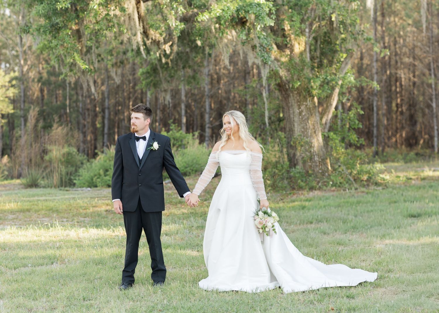 Bride and groom holding hands walking through green meadow with tall pine trees in background.