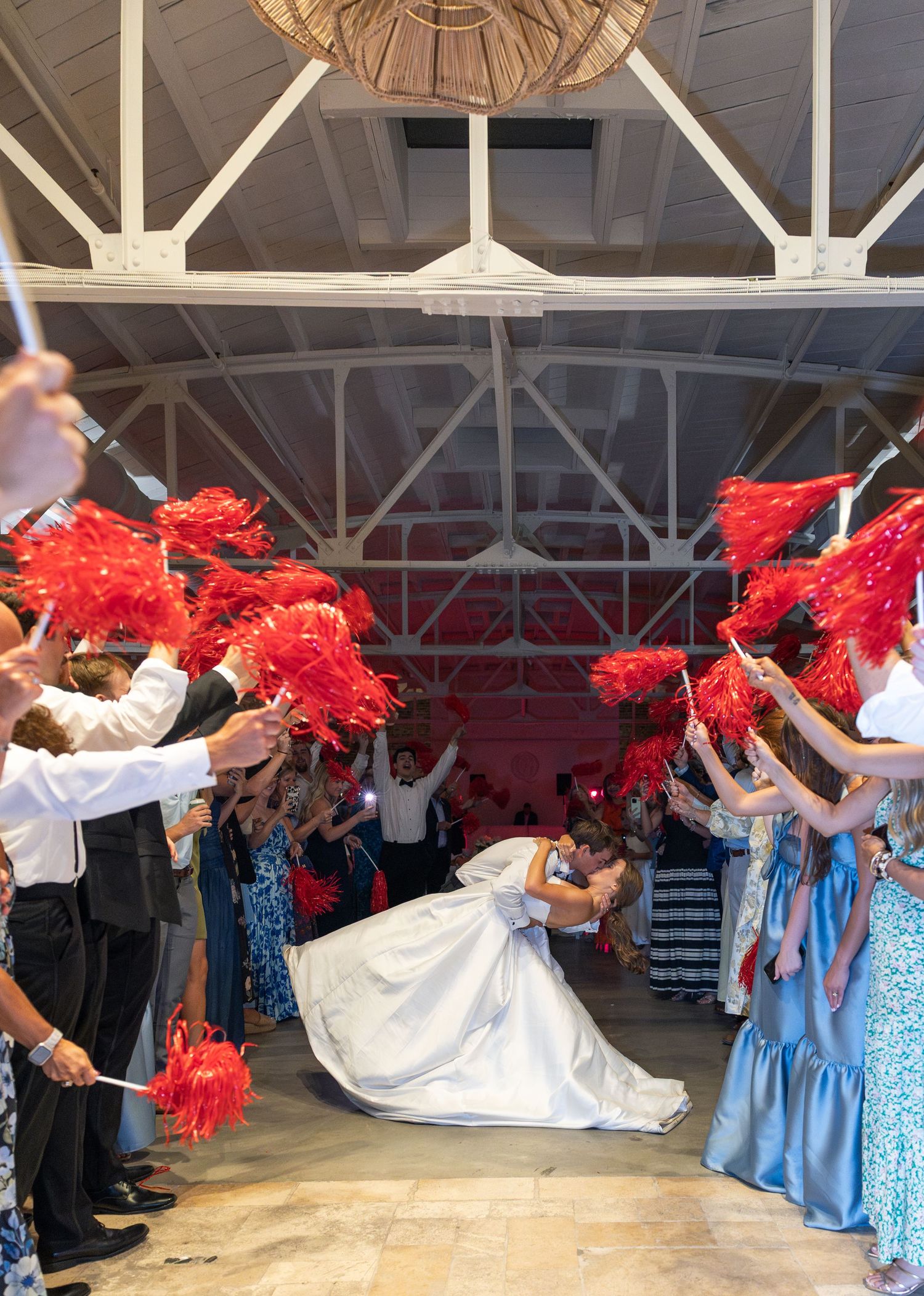 Bride and groom share dip kiss during grand exit with guests waving red pom-poms.