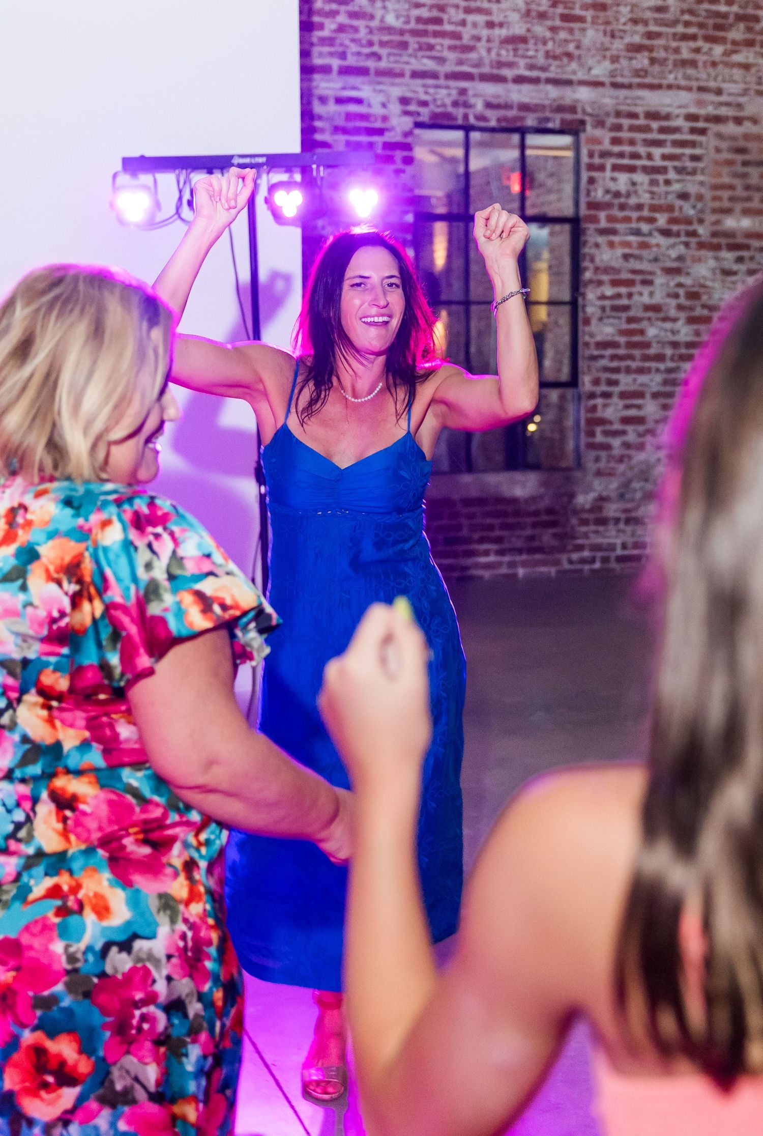 Guest in blue dress dances energetically under colorful lights at wedding reception.