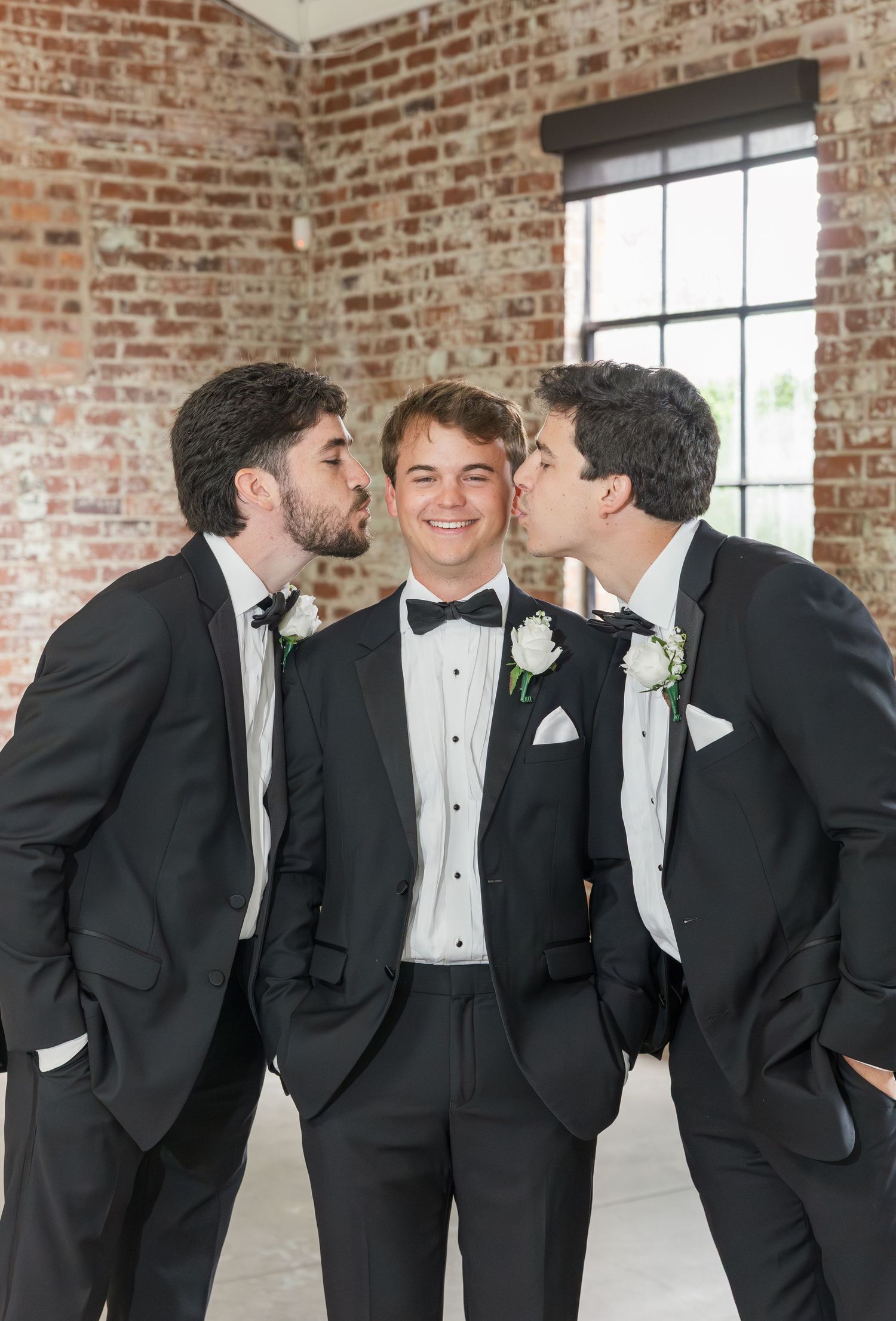 Two groomsmen kiss the groom on the cheeks in a fun wedding party portrait.