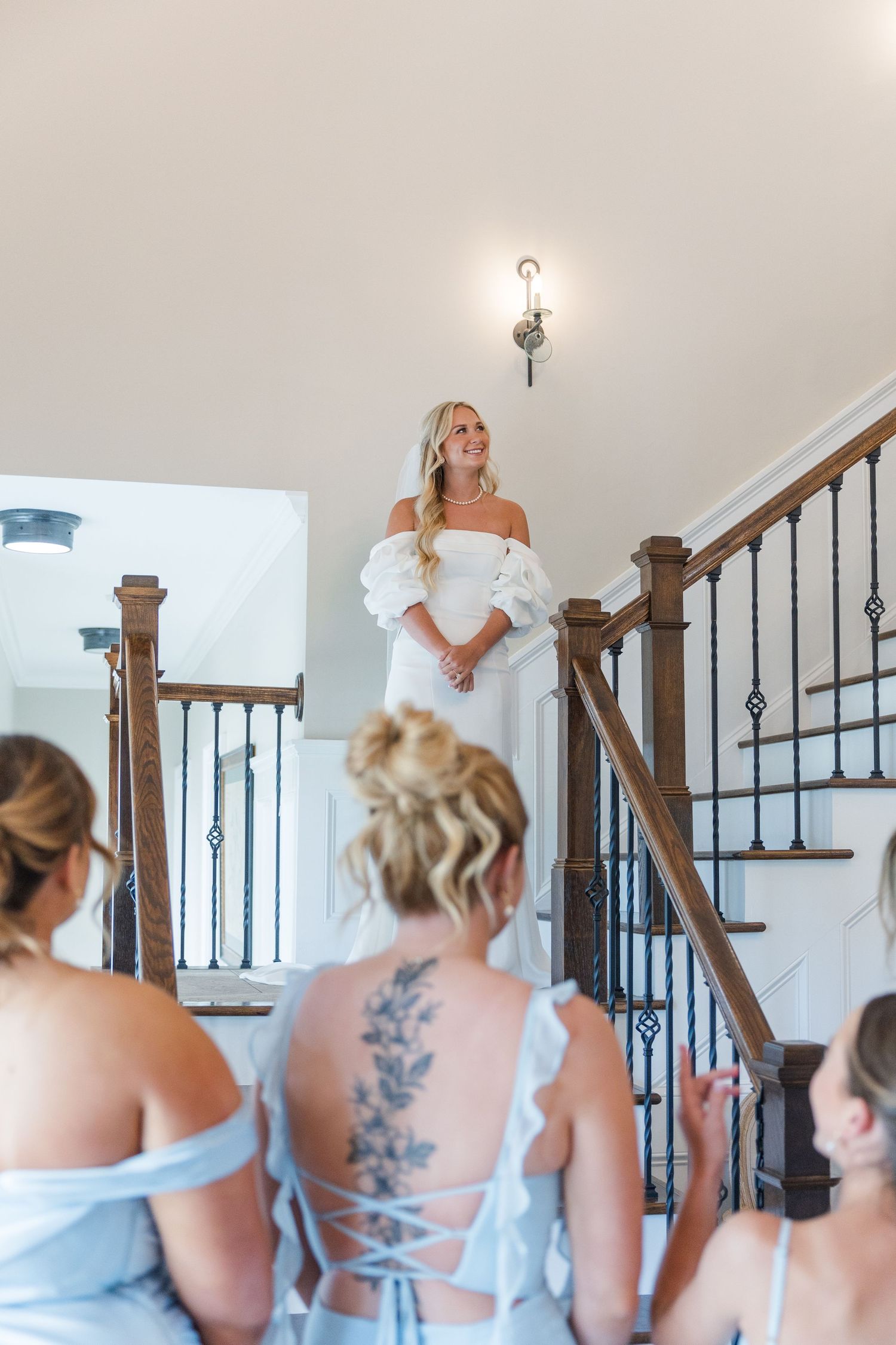 Bride in off-shoulder white gown descends staircase as bridesmaids in blue dresses look on admiringly.