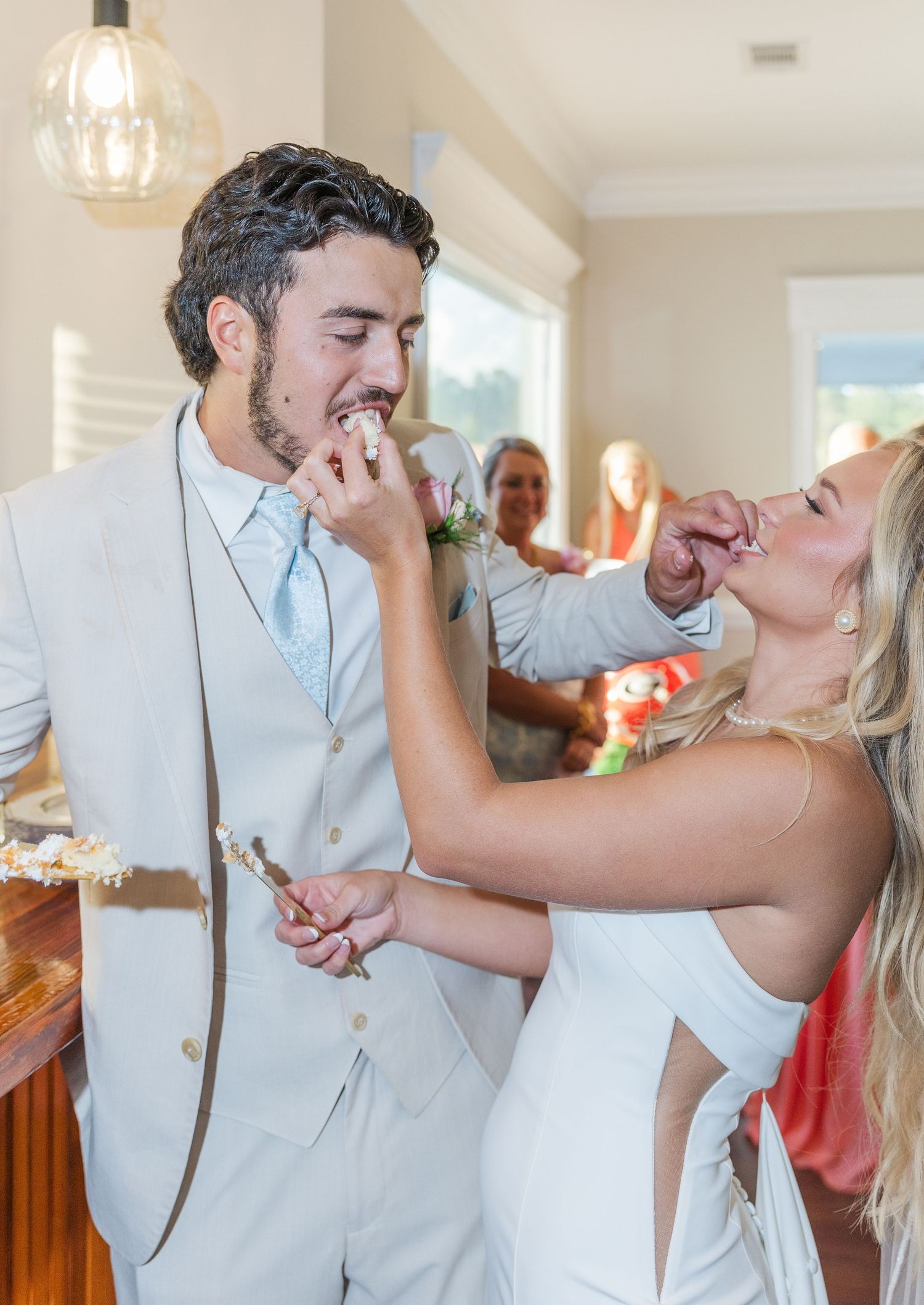 Bride and groom laugh while feeding each other wedding cake at colorful indoor reception celebration.