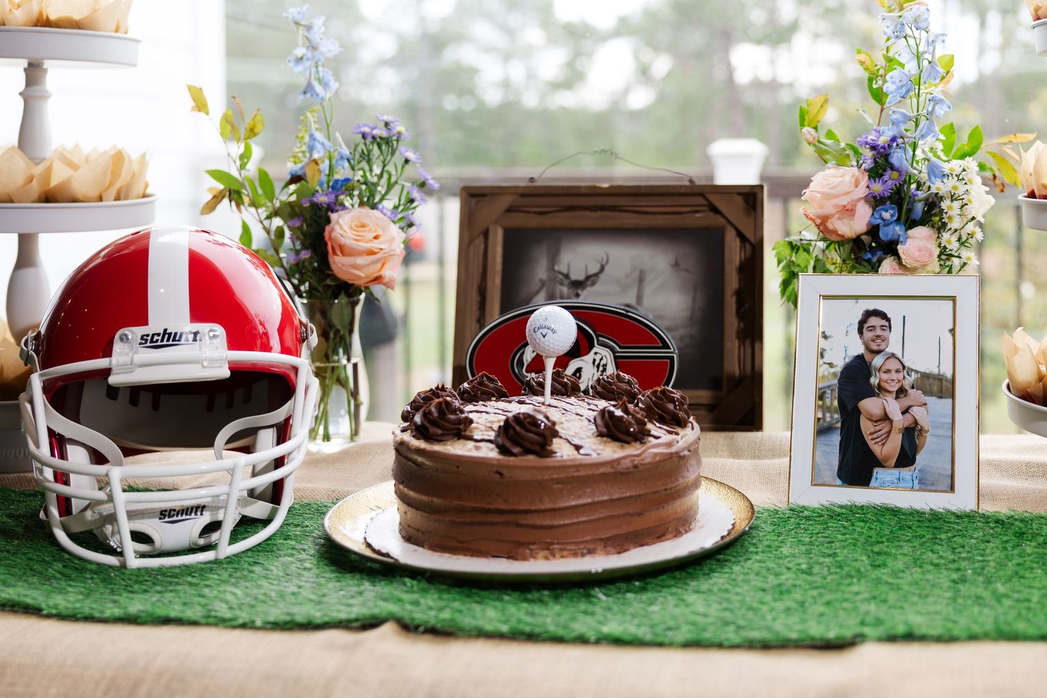 Grooms cake table featuring chocolate cake, football helmet, golf topper, and framed photo with floral accents.