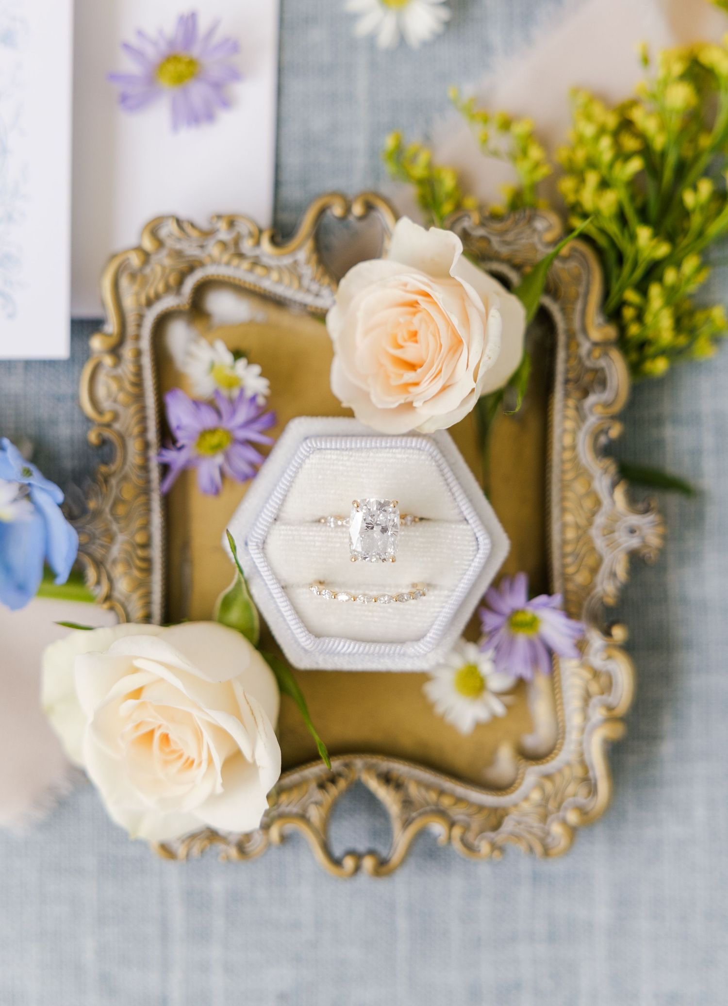 Elegant wedding rings displayed in a hexagonal velvet box surrounded by cream roses and purple wildflowers on a gold ornate tray.