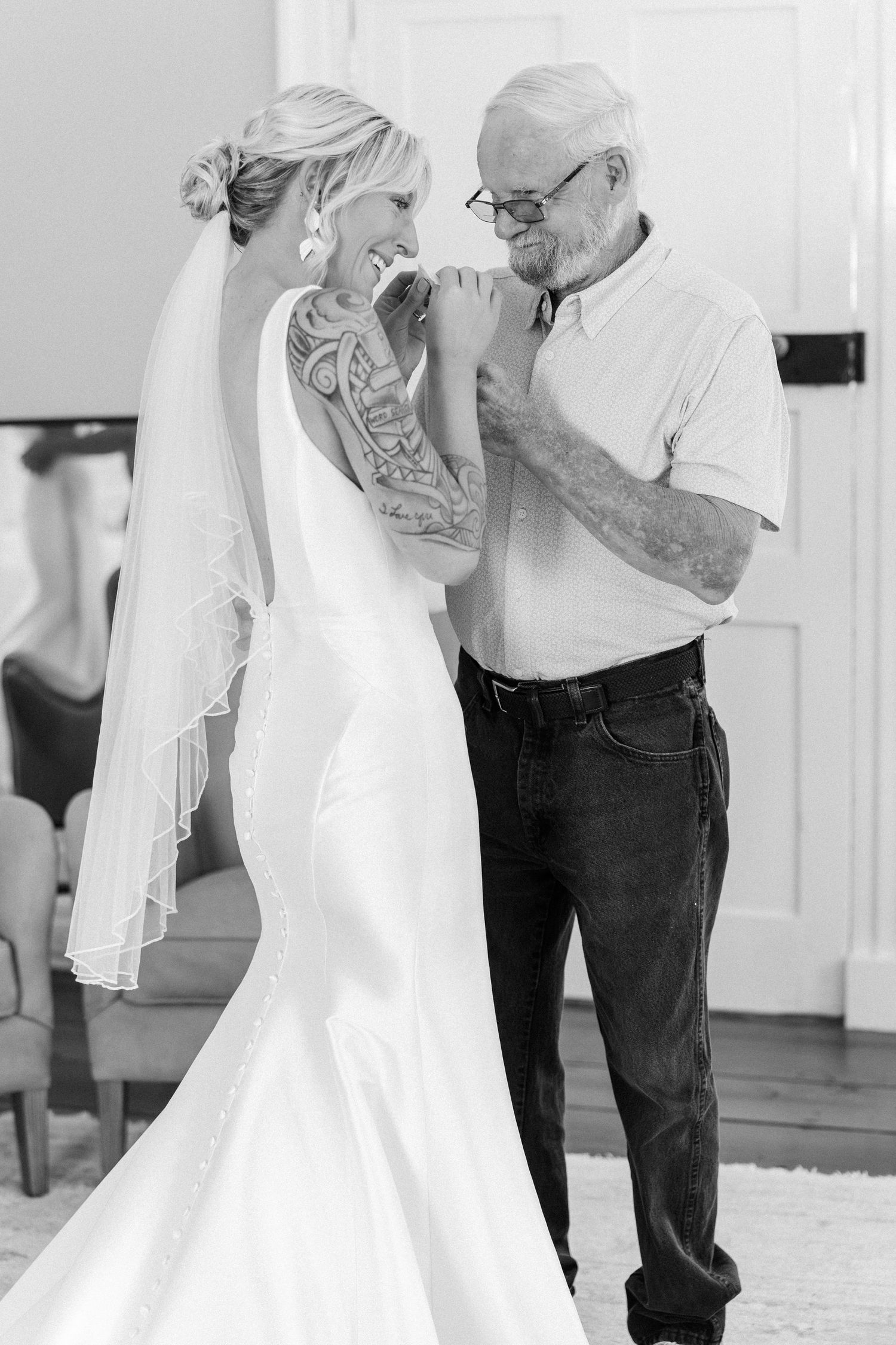 Black and white photo of tattooed bride in wedding gown sharing tender moment with older gentleman.