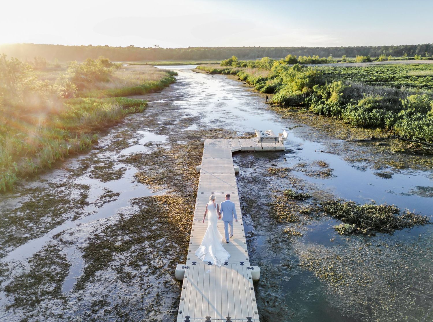 Aerial view of a couple holding hands on a wooden boardwalk over a coastal marsh waterway at sunset.
