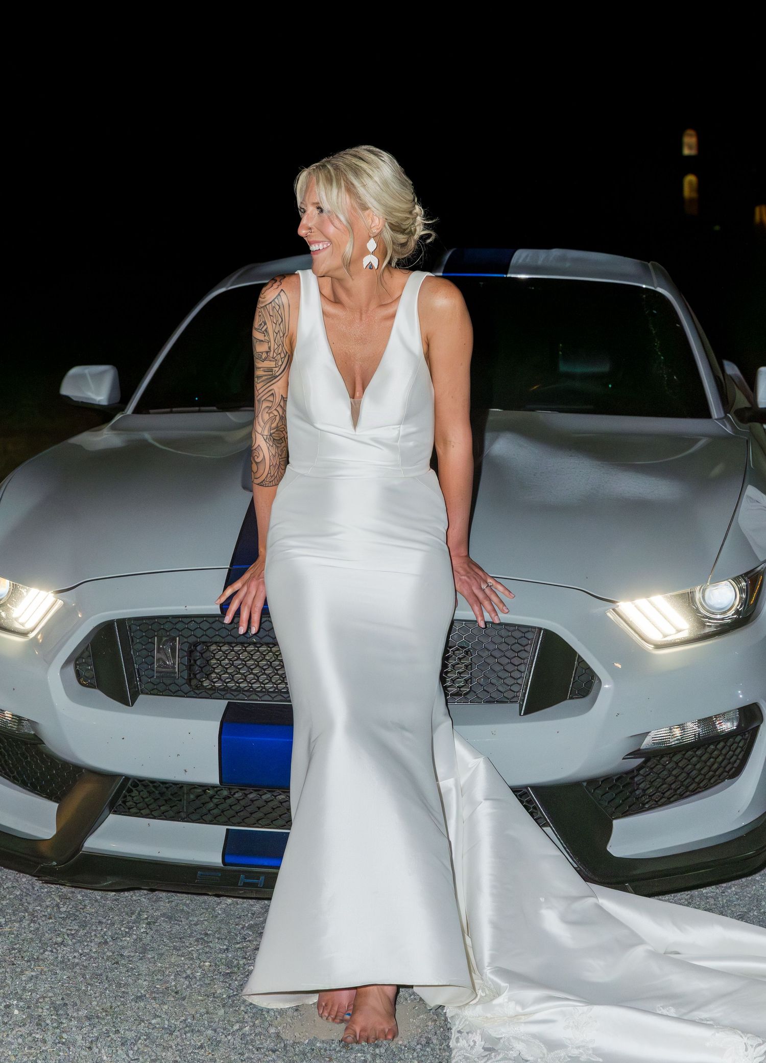 Tattooed bride in sleek white mermaid gown poses against silver Ford Mustang GT350 at night.