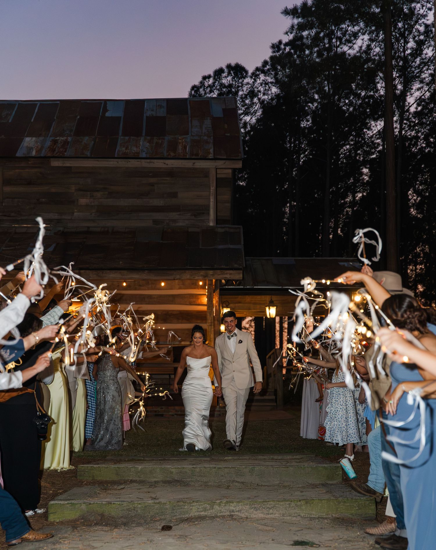 Bride and groom exit rustic barn venue through cheering guests waving ribbon wands at twilight.