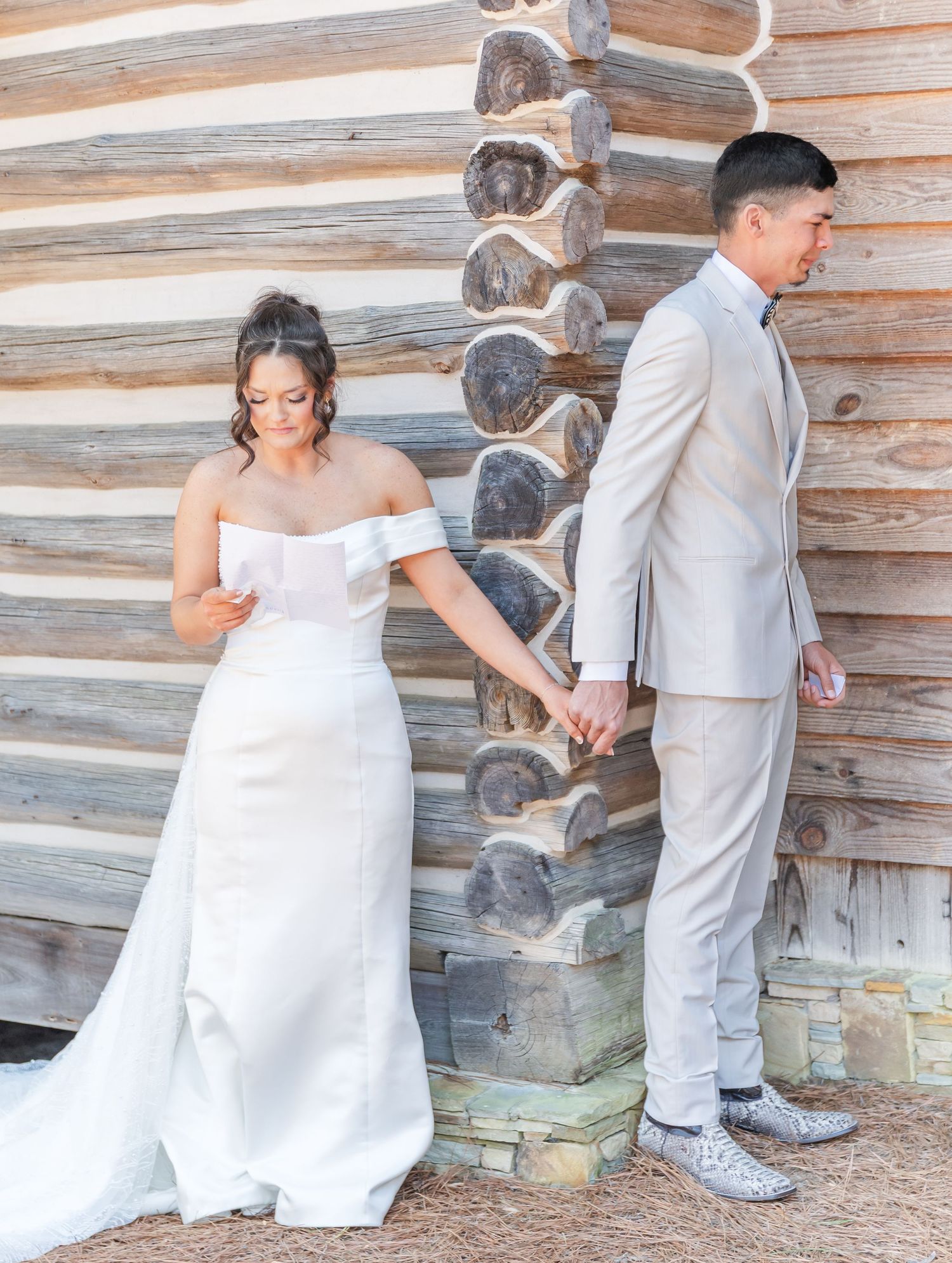 Bride and groom share a first look moment holding hands against a rustic log cabin wall before their wedding ceremony.