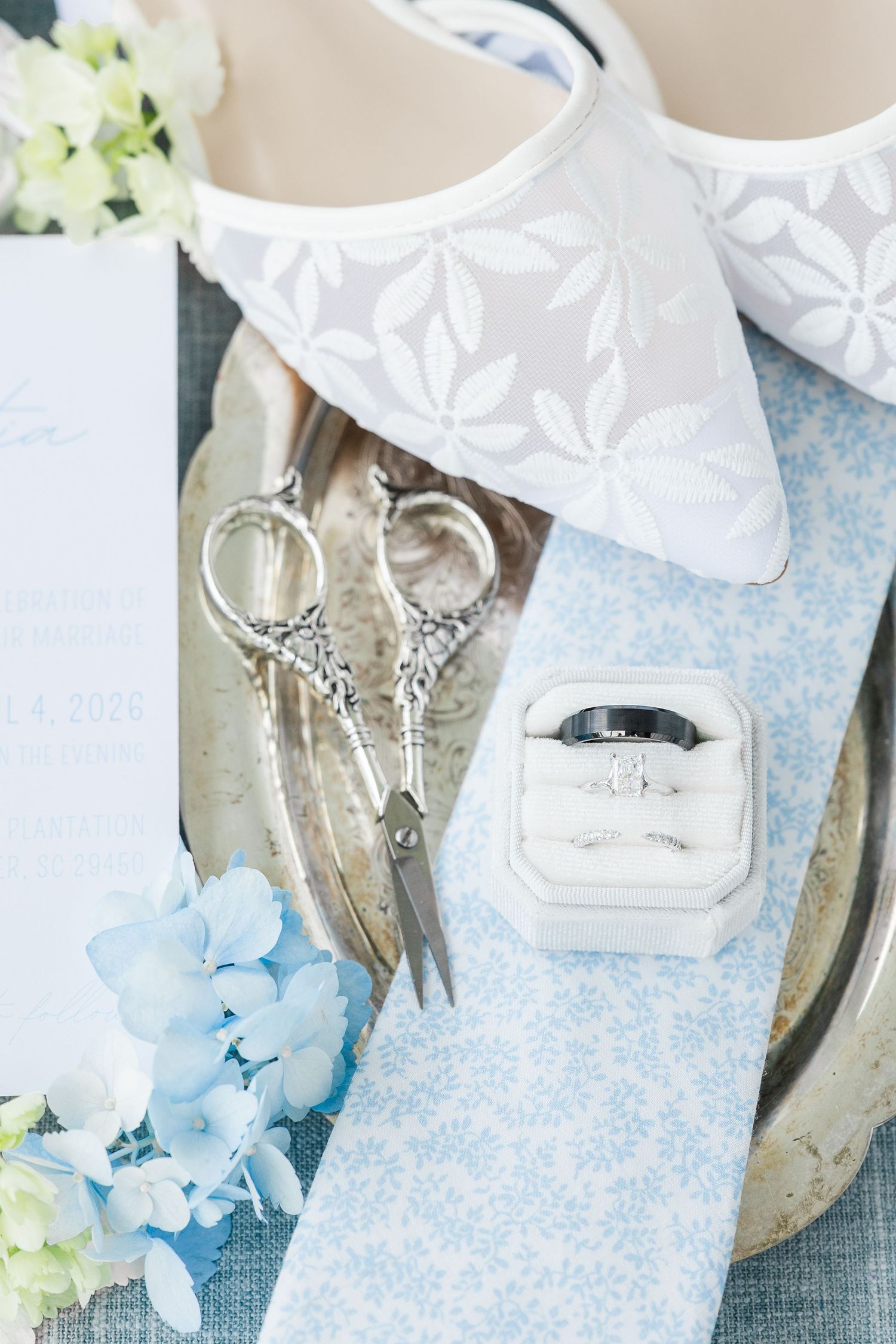 Elegant wedding flat lay with white lace heels, diamond rings in box, ornate scissors, blue hydrangeas, and invitation on blue fabric.