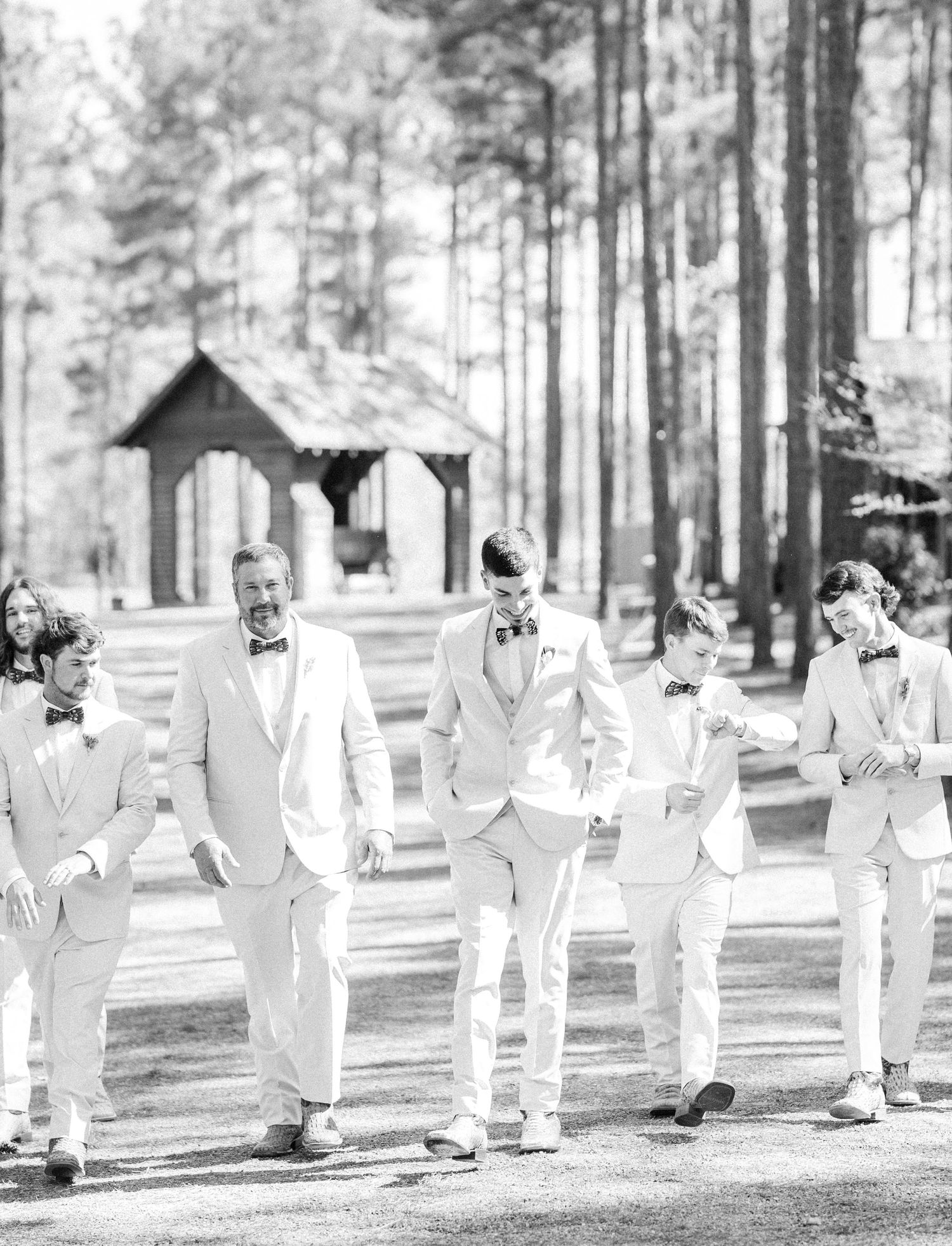 Groomsmen in cream suits walk together through a pine forest wedding venue in black and white photo.