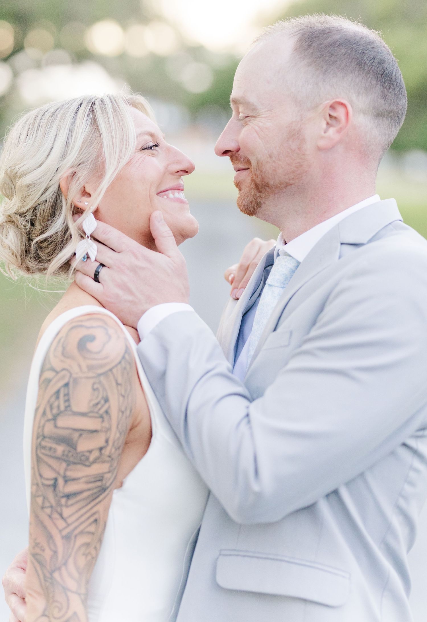 Smiling bride with arm tattoo and groom in light blue suit share a tender moment at their outdoor wedding.
