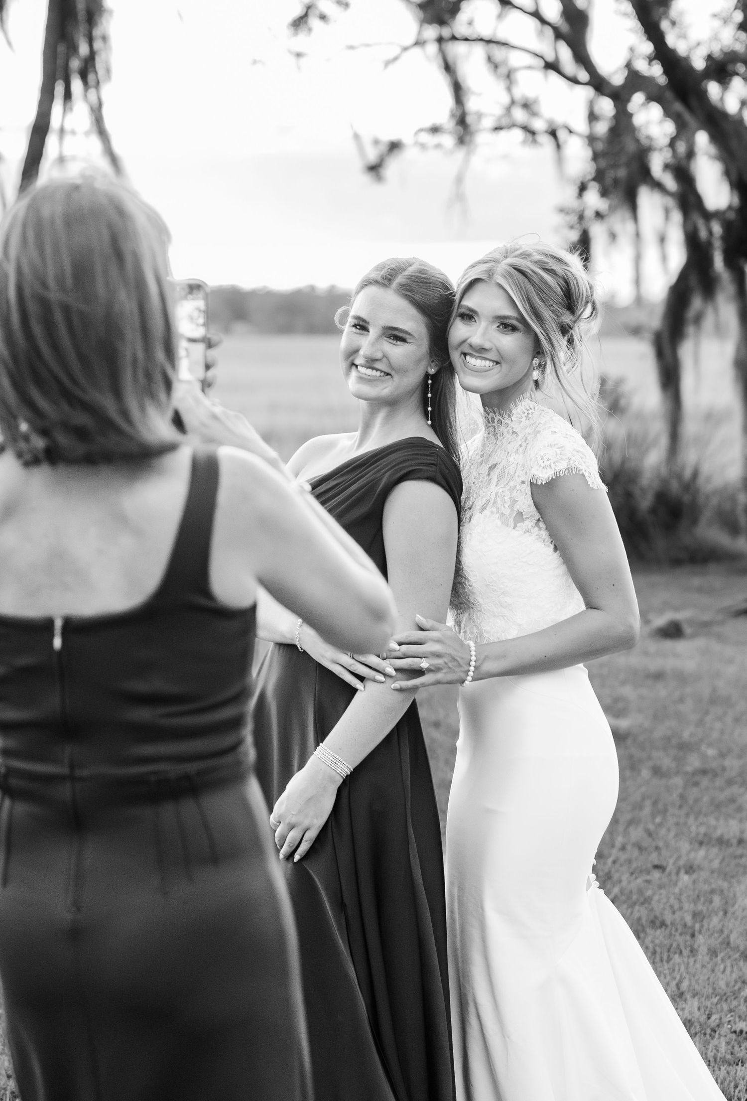 Black and white photo of a bride in lace gown posing with bridesmaid in dark dress outdoors at wedding.