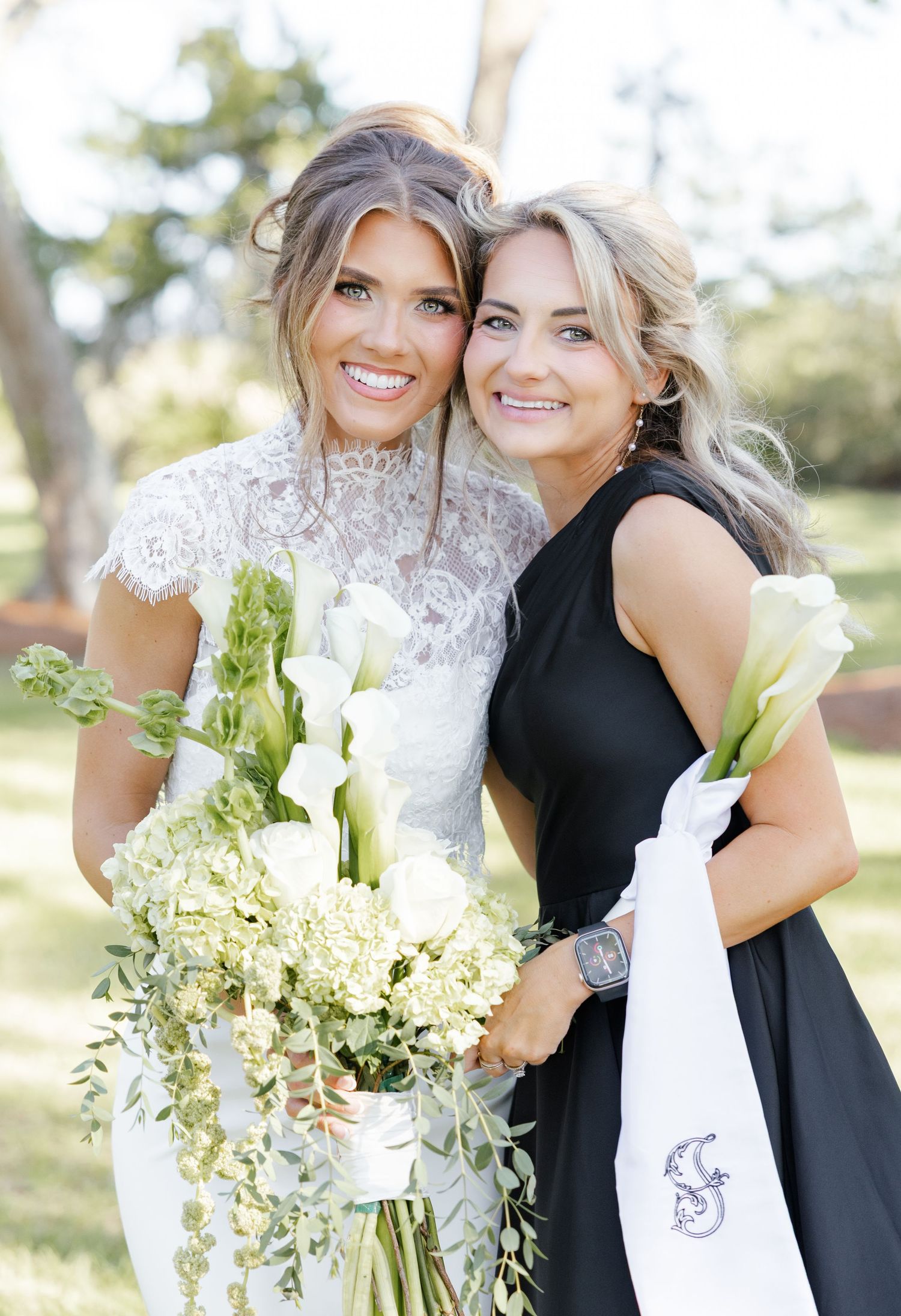 Smiling bride in lace gown holds white calla lily bouquet beside bridesmaid in black dress outdoors.