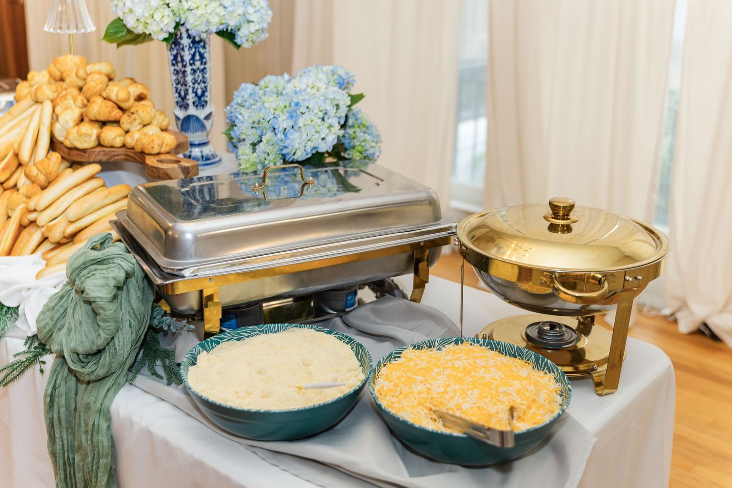 Wedding reception buffet table with chafing dishes, creamy dips, bread rolls, and blue hydrangea centerpiece.