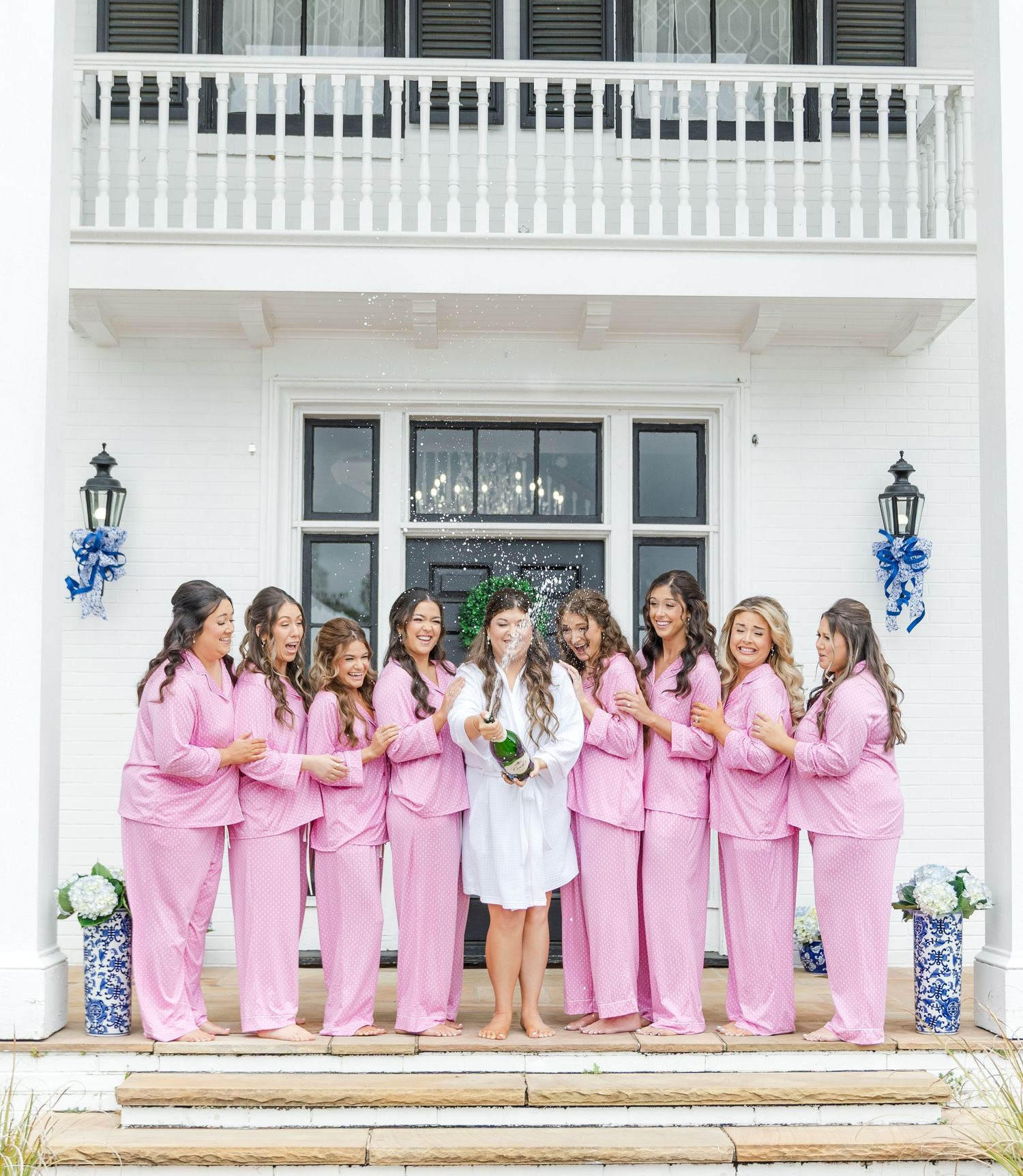 Bride in white robe poses with bridesmaids in matching pink pajamas on steps of white venue.