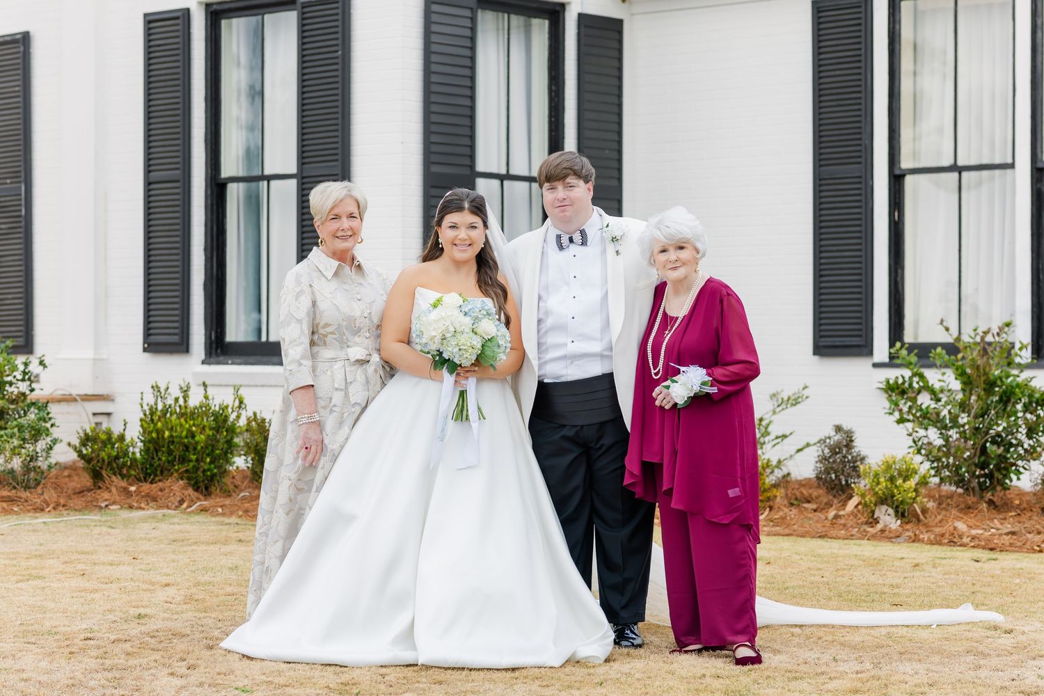 Bride and groom pose with two grandmothers in formal attire outside white wedding venue after ceremony.