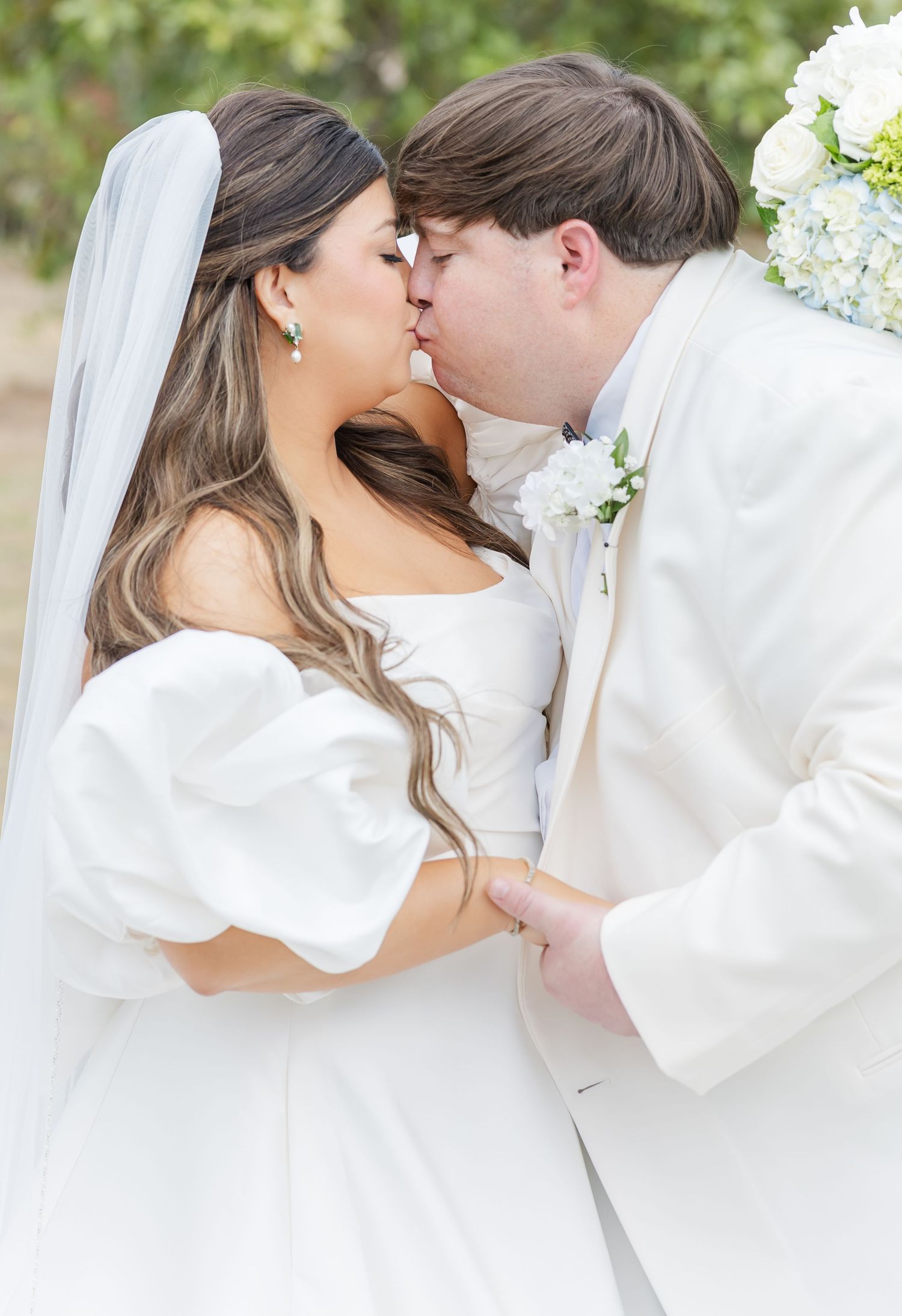 Bride and groom share a romantic kiss, she in a white puff-sleeve gown with veil, he in a white suit with floral boutonniere.