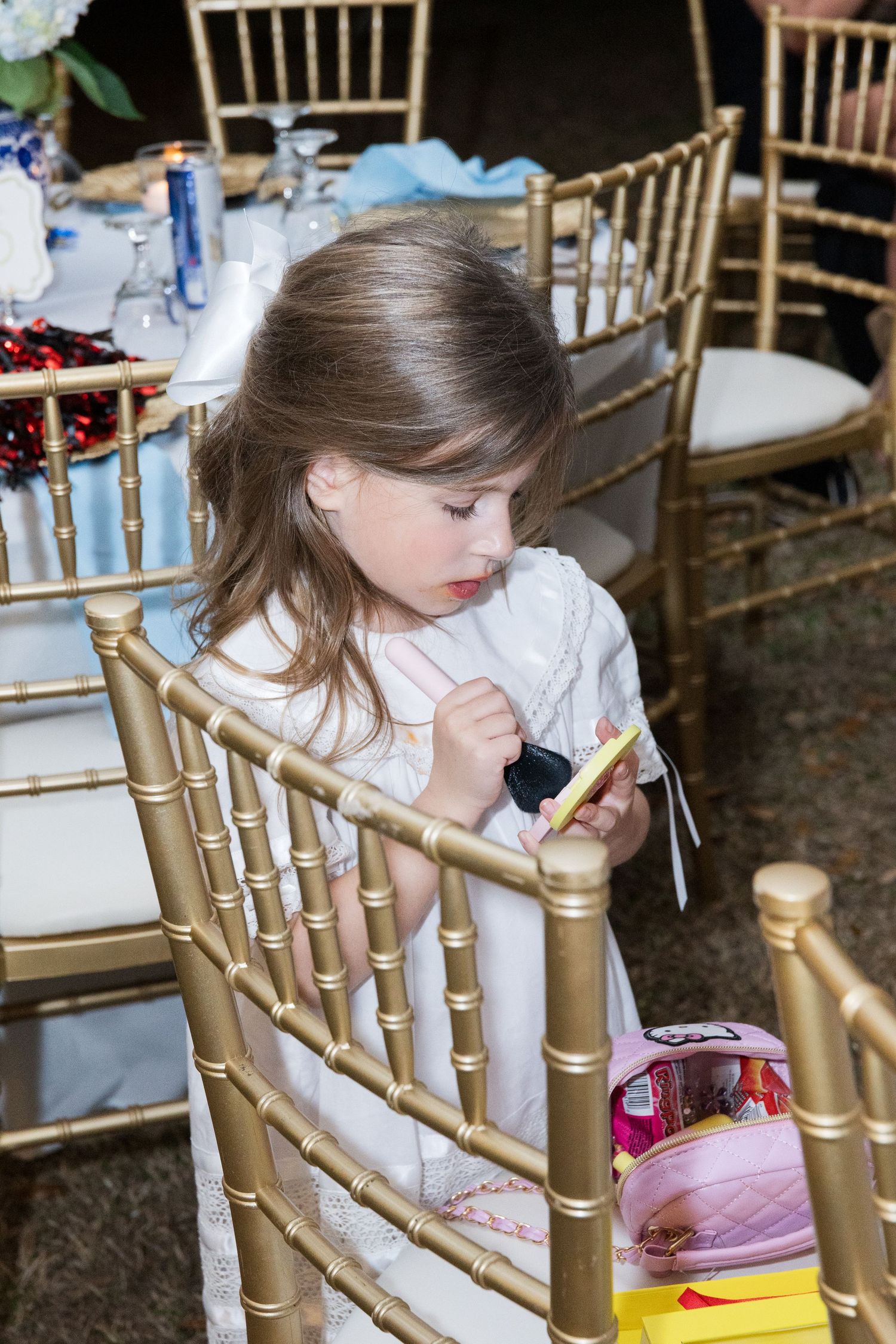 Young flower girl in white dress sitting on gold chiavari chair at wedding reception.