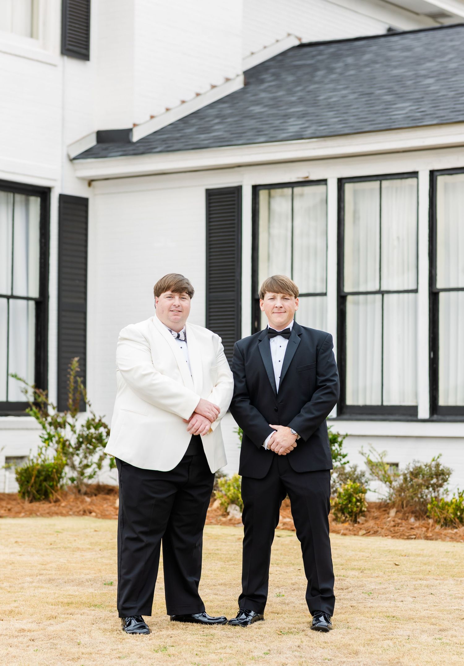 Groom in white tuxedo jacket and groomsman in black tuxedo posing outside white wedding venue.