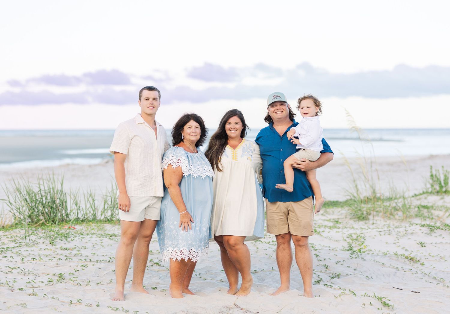 A group of people standing together on a sandy beach at sunset, with ocean and dunes in the background.