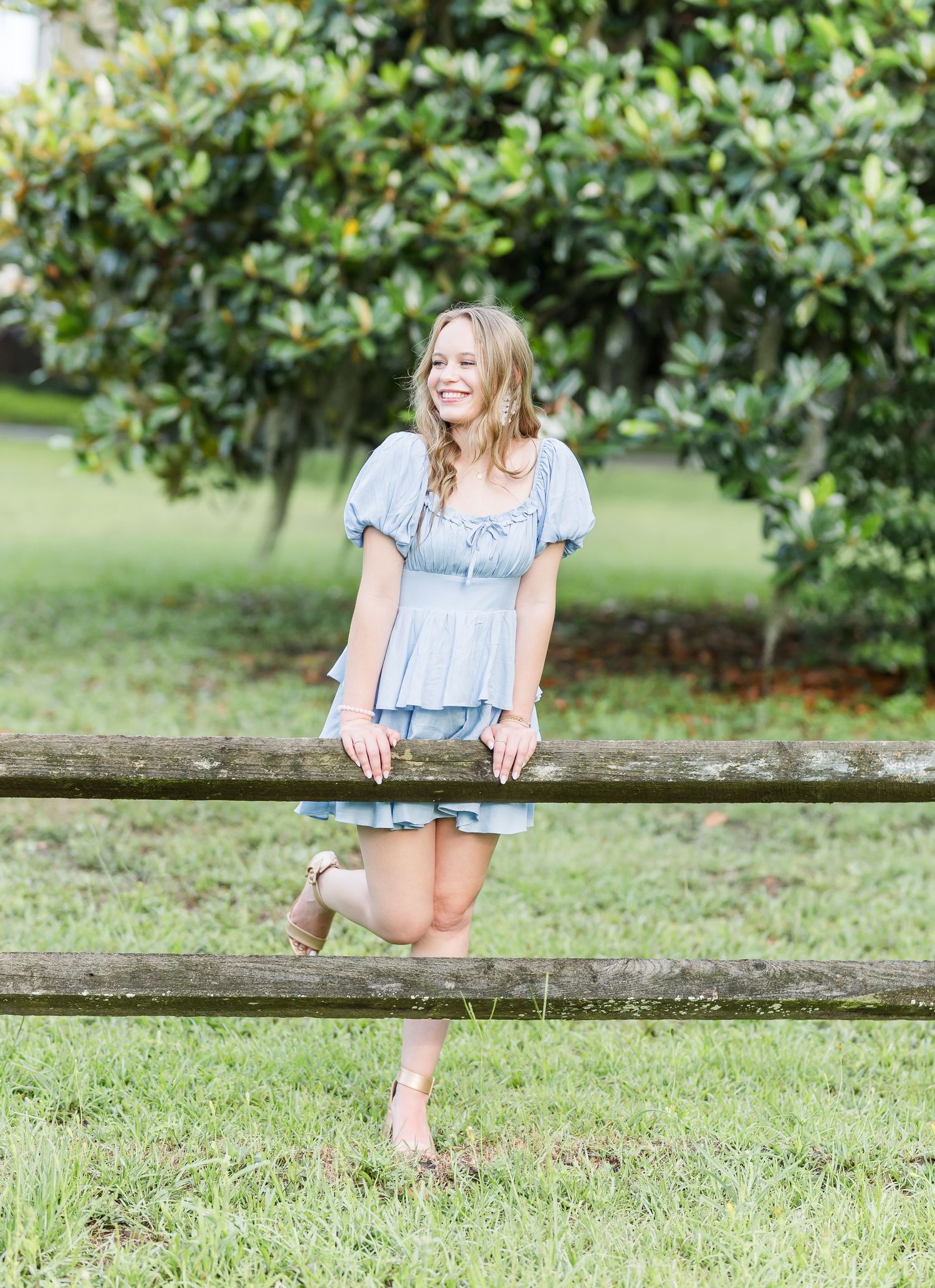 A girl in a blue dress sits casually on a wooden fence surrounded by lush greenery.