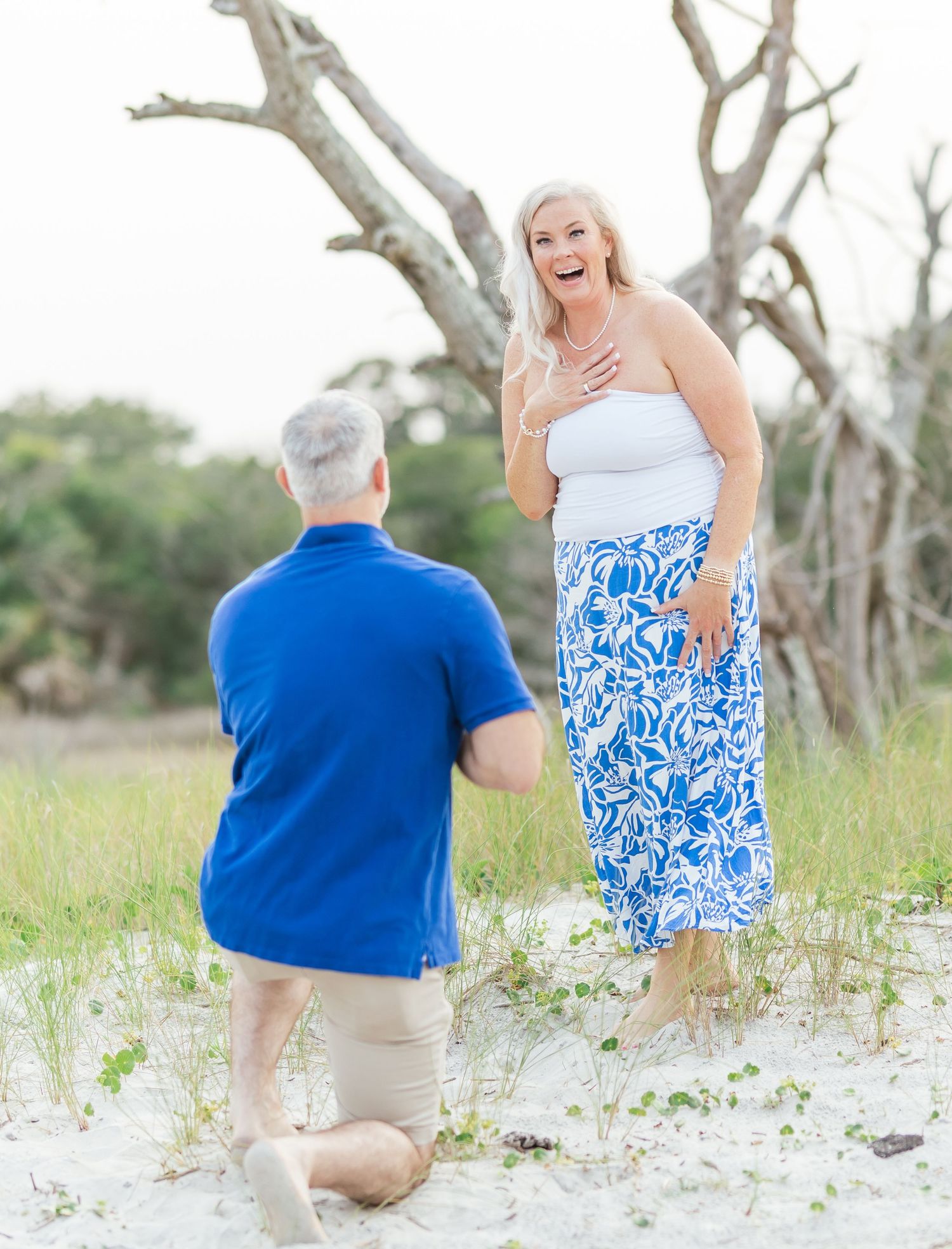 A surprise marriage proposal moment on a beach with bare trees and green foliage in the background.
