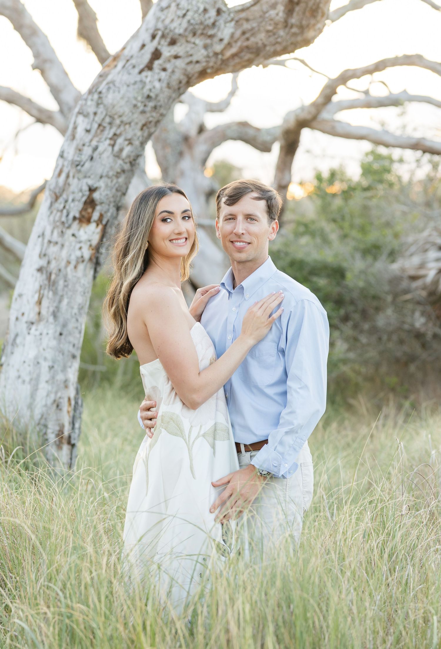 Smiling couple standing in tall grass field embracing near a white birch tree at sunset.