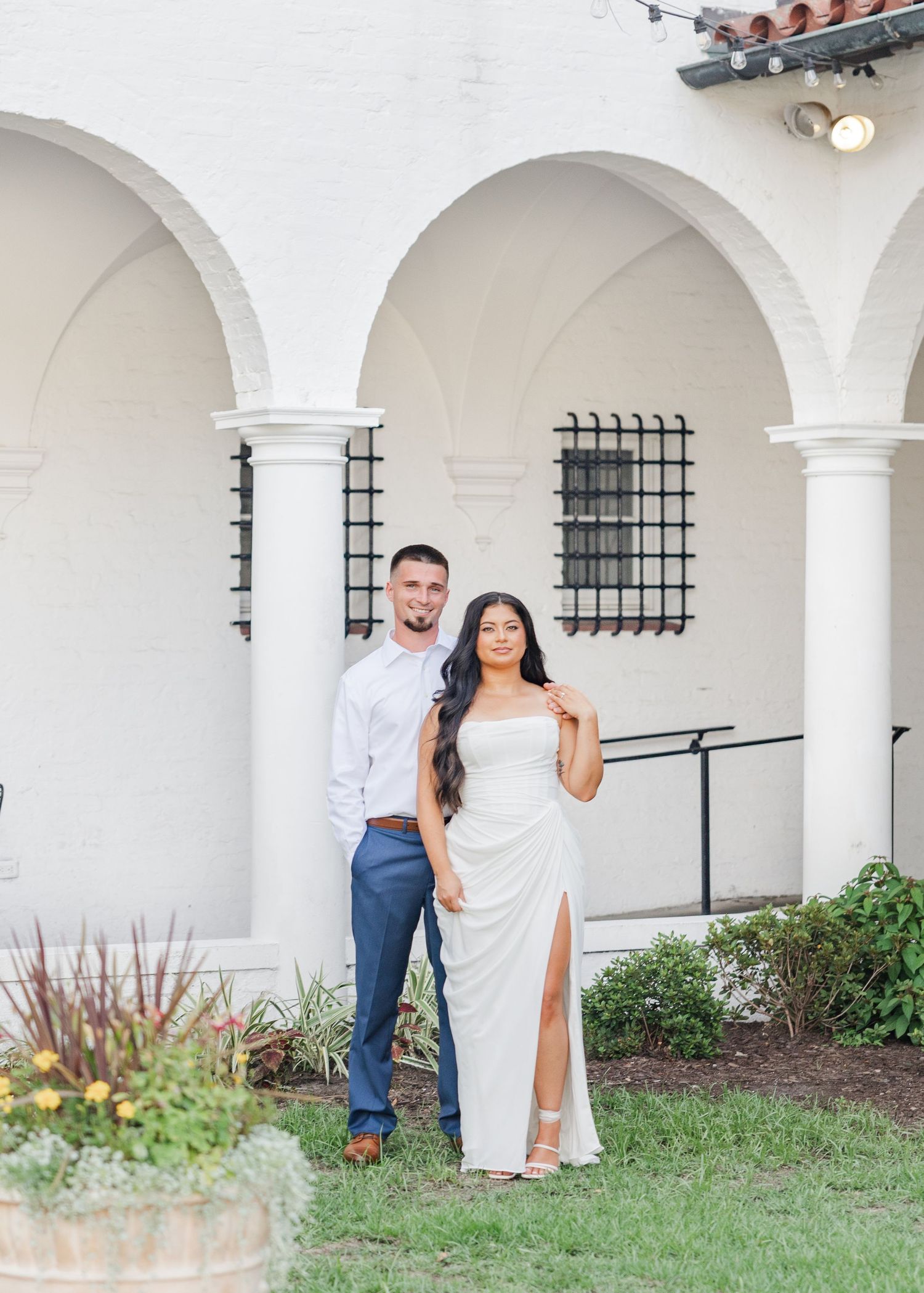 Elegant couple posing under white arched colonnade, woman in white slit gown and man in blue suit at outdoor venue.