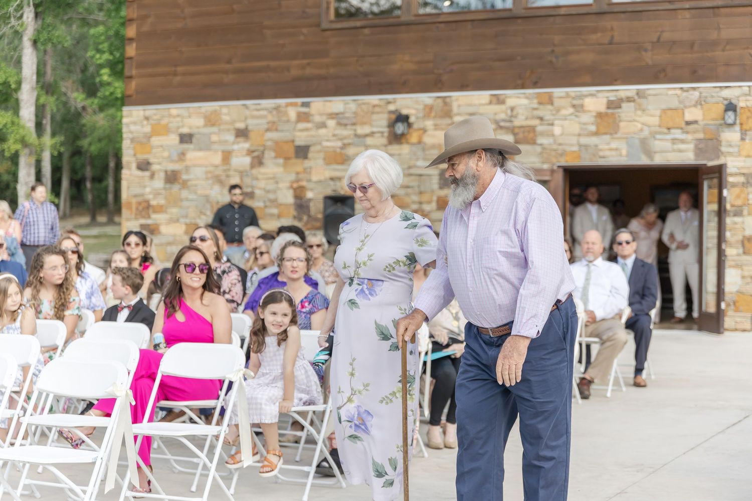 Wedding ceremony procession outside rustic stone building with guests watching.