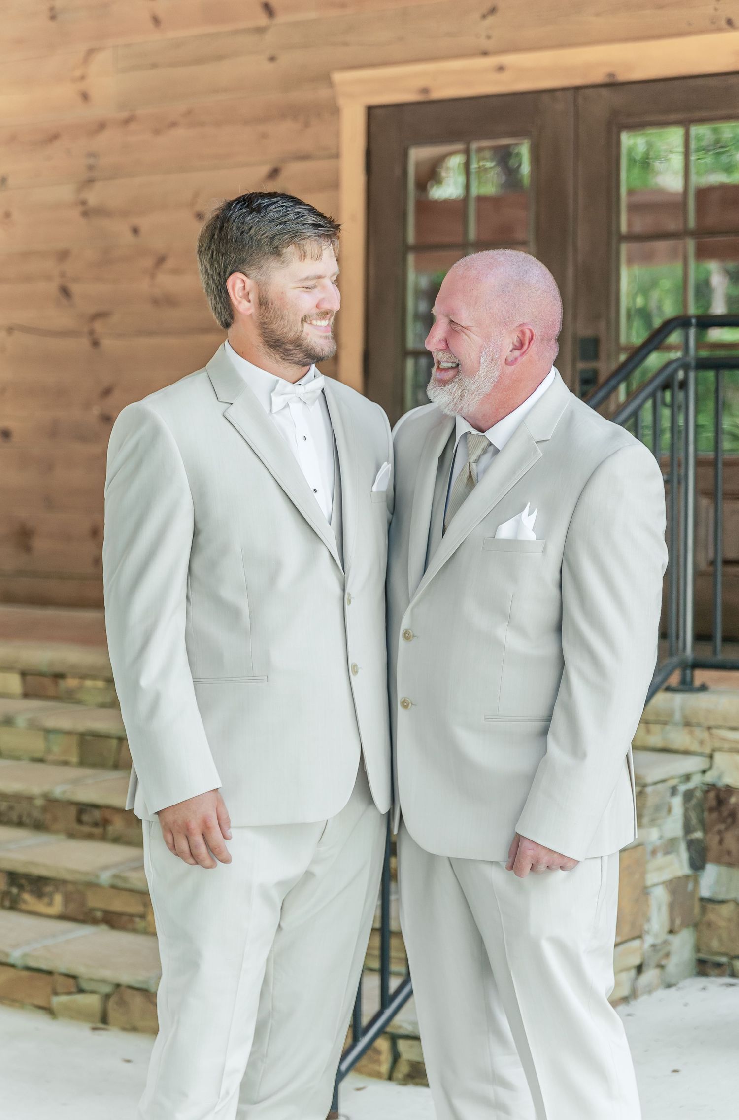 Two people in light beige suits share a joyful moment on a rustic wooden porch during a wedding celebration.