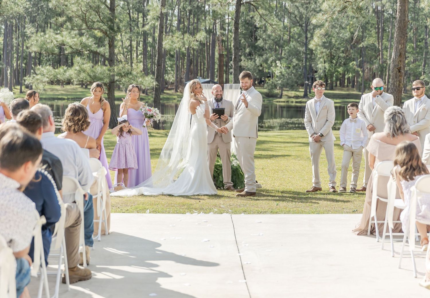 Scenic outdoor wedding ceremony with guests seated on white chairs and pine trees in background.