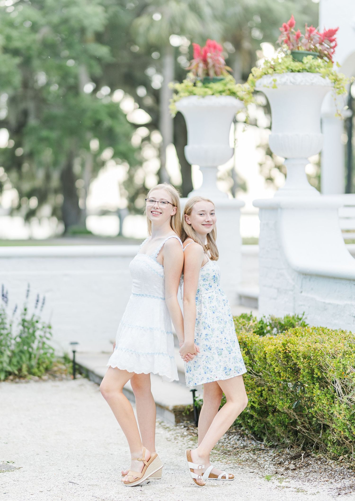 Two young women in light summer dresses pose together by decorative garden urns with flowers.