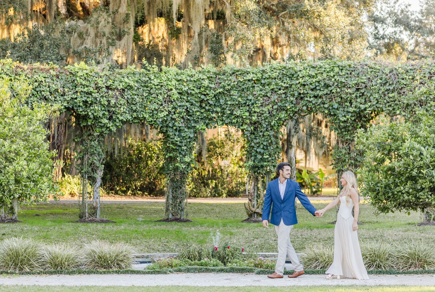 Two people walk together under a romantic archway covered in lush green ivy at a scenic outdoor location.