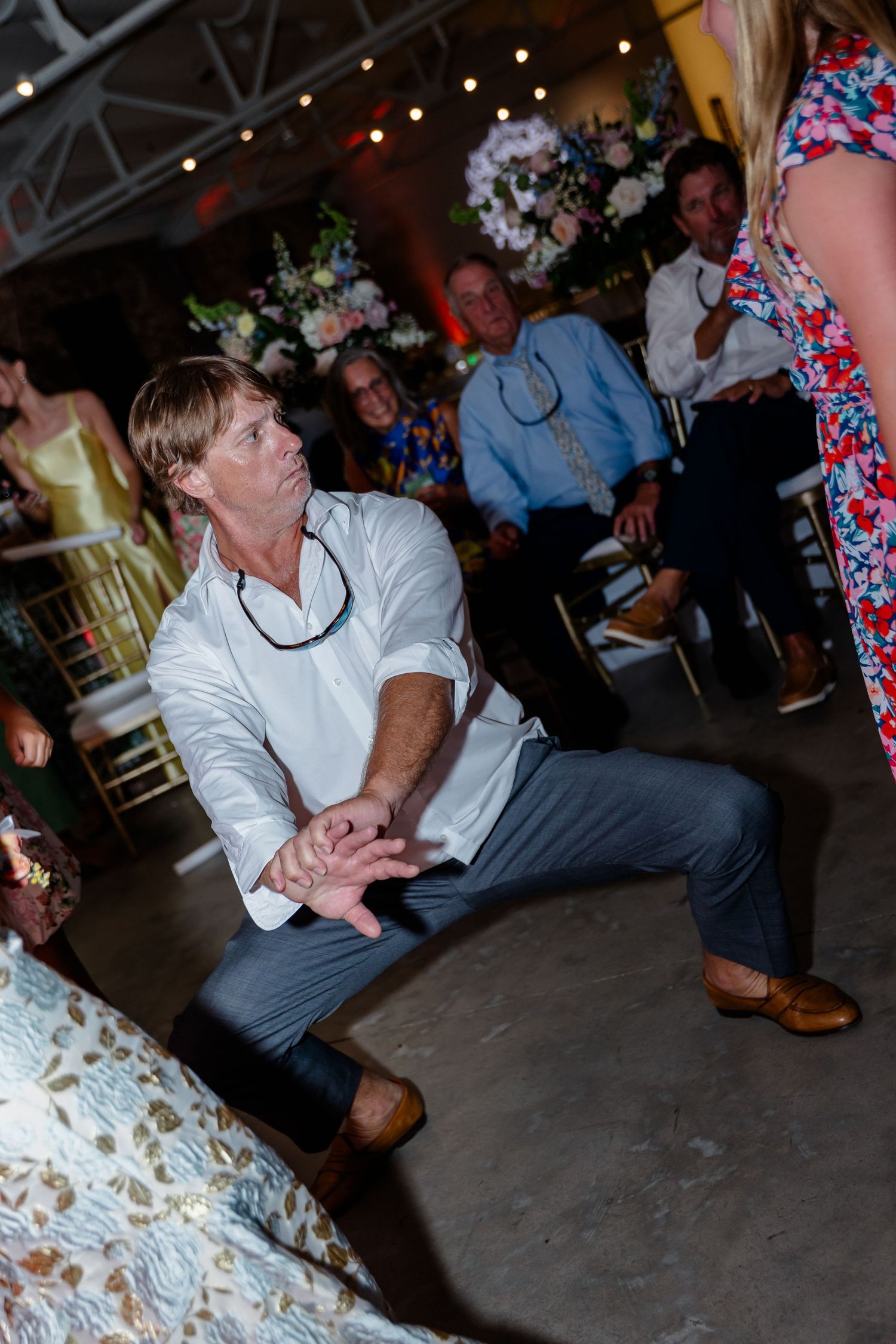 Wedding guest in white shirt dancing energetically at reception with floral arrangements and string lights overhead.