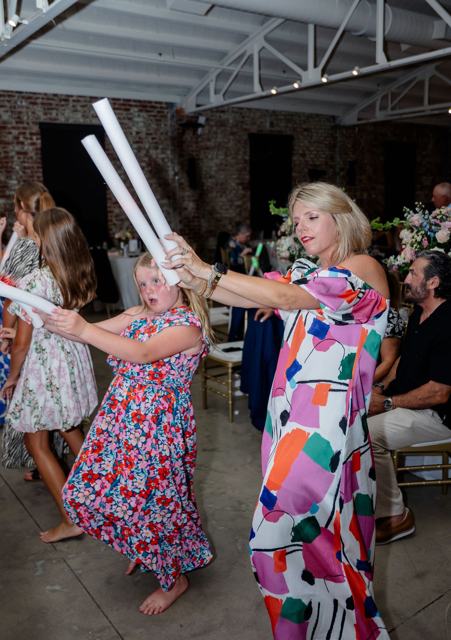 Wedding guests dancing with light-up foam sticks at a reception in an industrial brick venue.