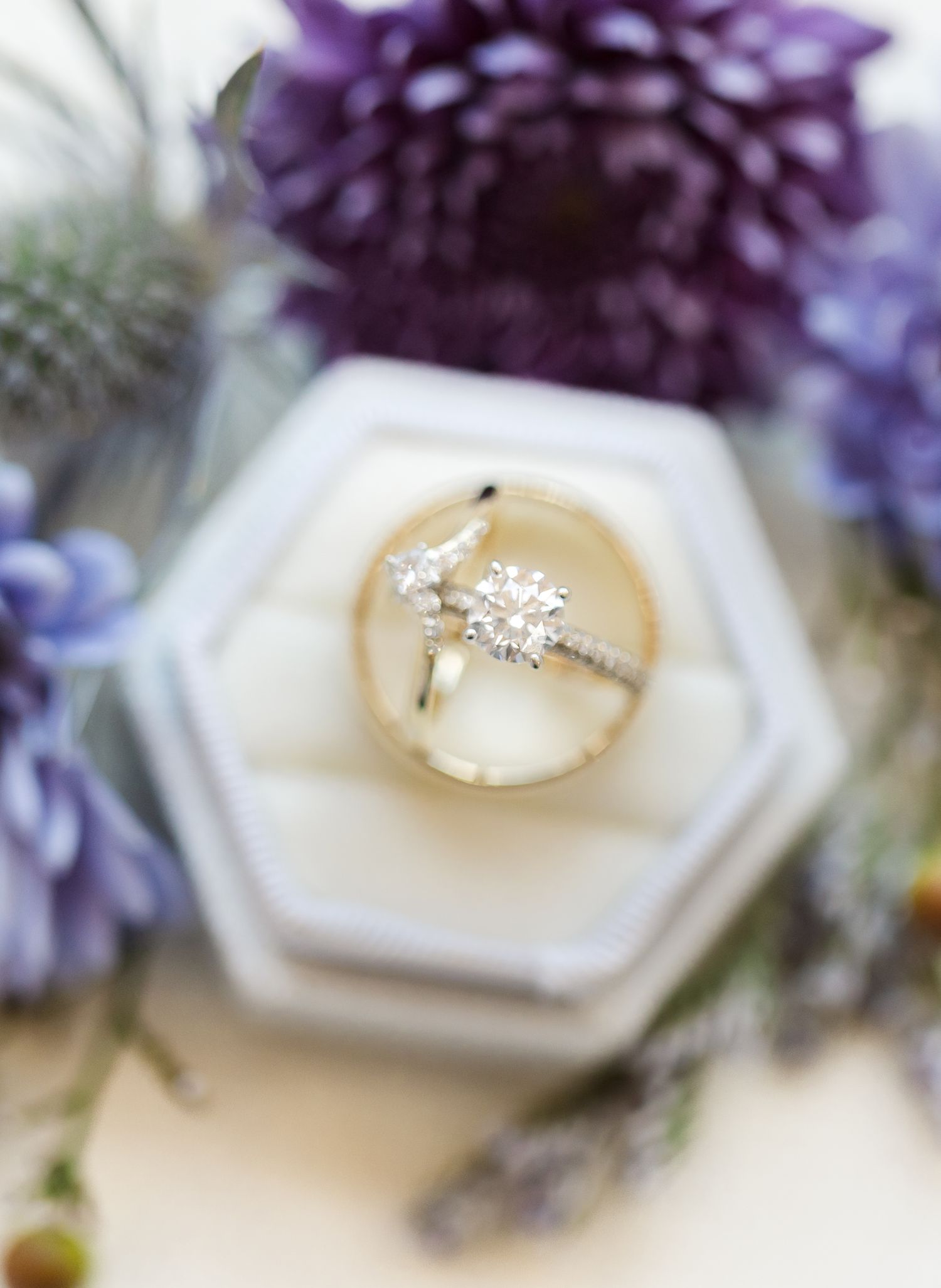 Elegant diamond engagement ring with purple flowers and white hexagonal ring box in soft focus background.