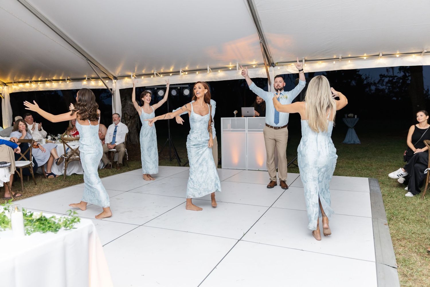 Wedding party dancing in a line formation on a white dance floor under an outdoor reception tent.