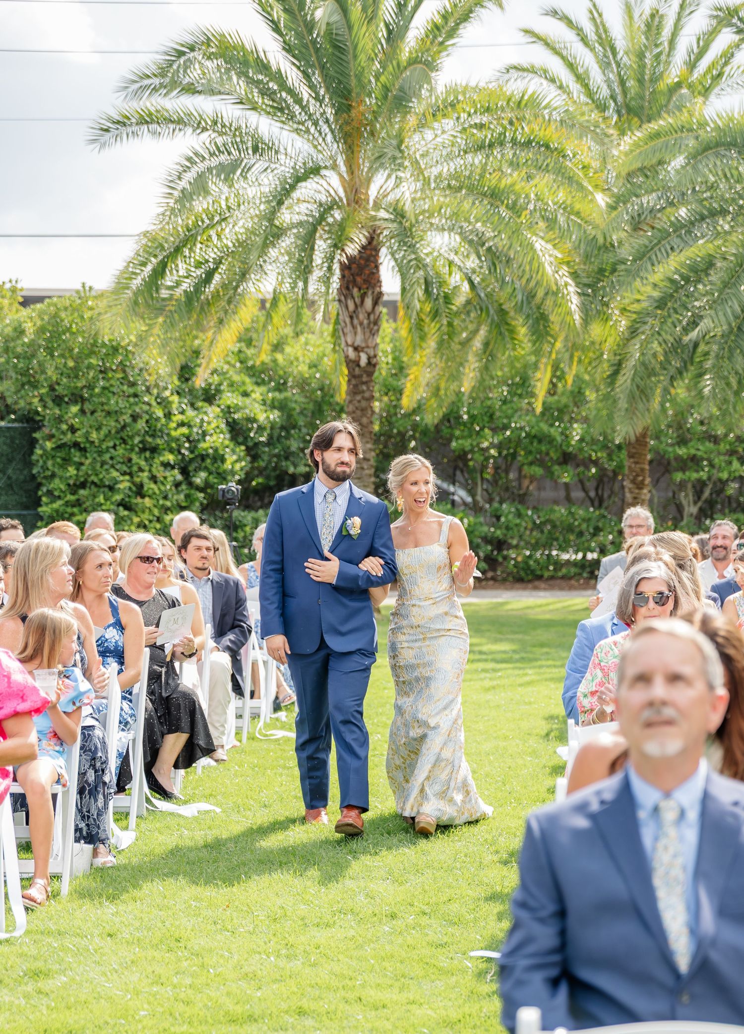 Wedding guests gather on a lush green lawn surrounded by tropical palm trees during an outdoor ceremony.