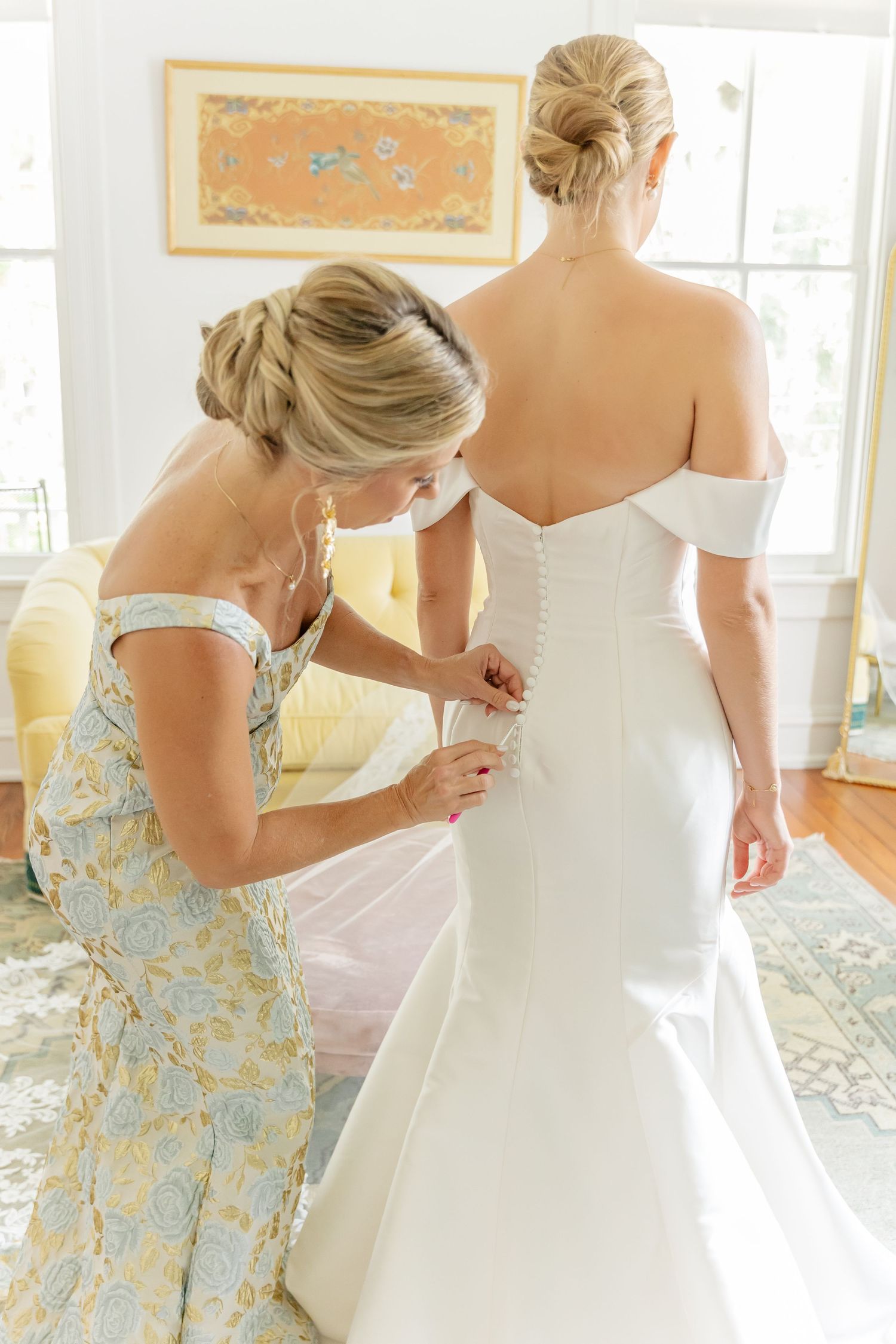 A bride in a fitted white off-shoulder wedding gown gets help with final dress adjustments in a sunlit room.