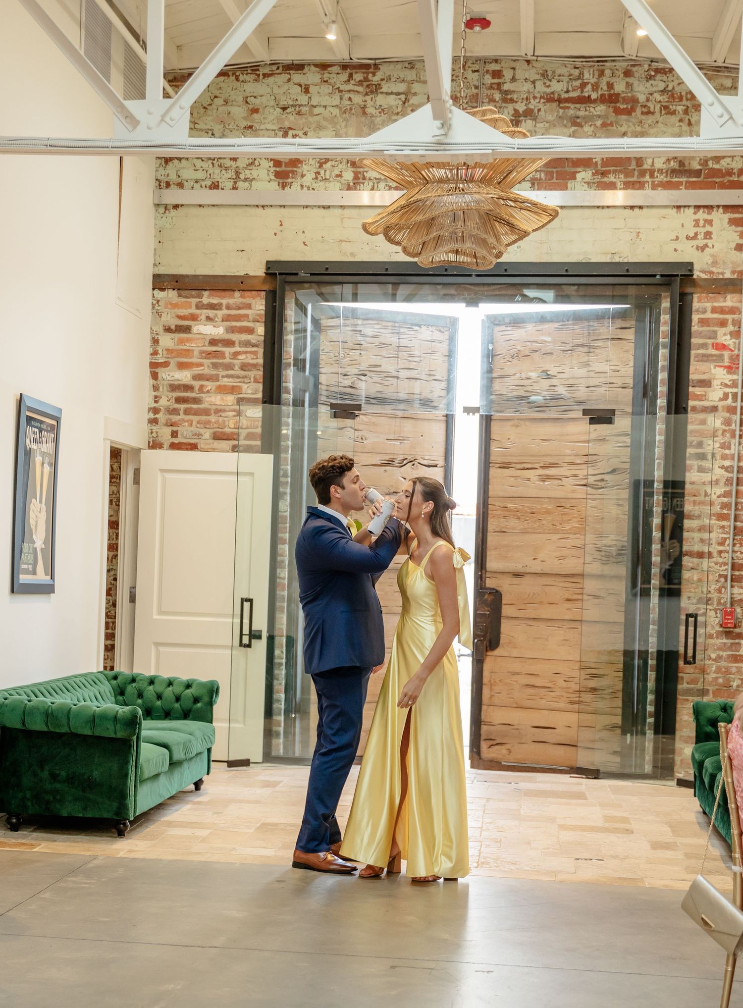 A couple shares an intimate moment in a rustic brick-walled venue with emerald velvet couches and wooden accents.