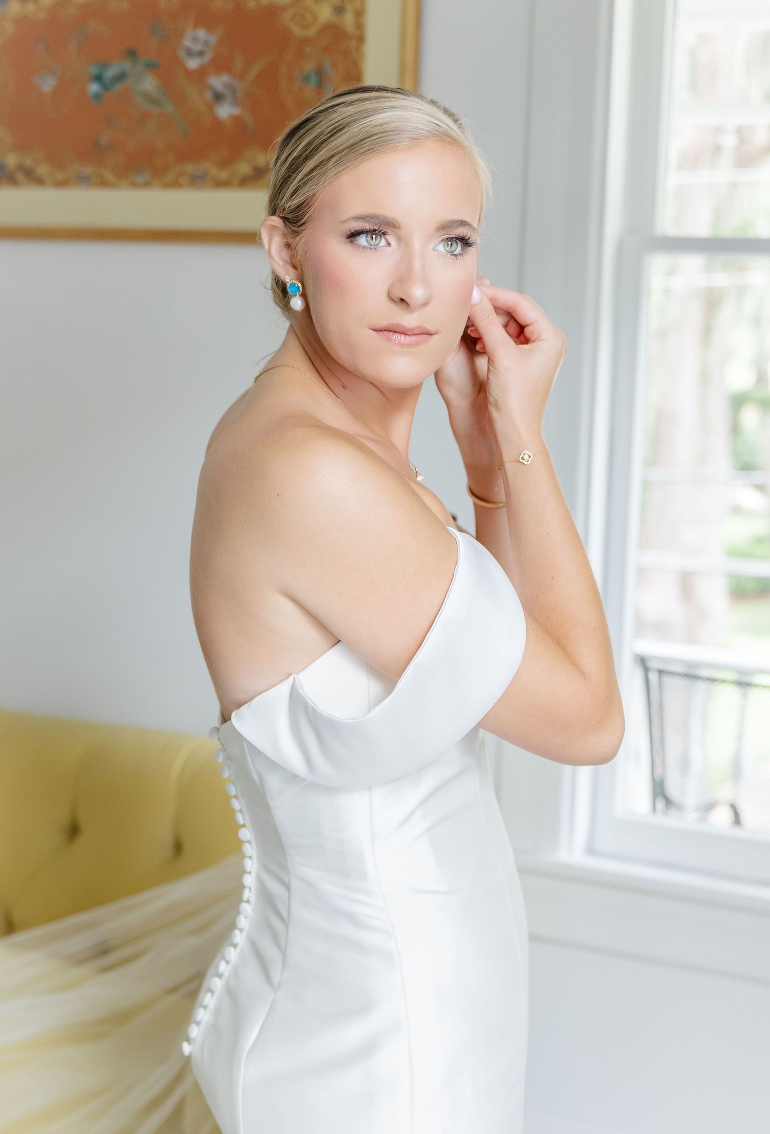 Elegant bride in a classic white one-shoulder wedding gown poses gracefully by a bright window in a light-filled room.