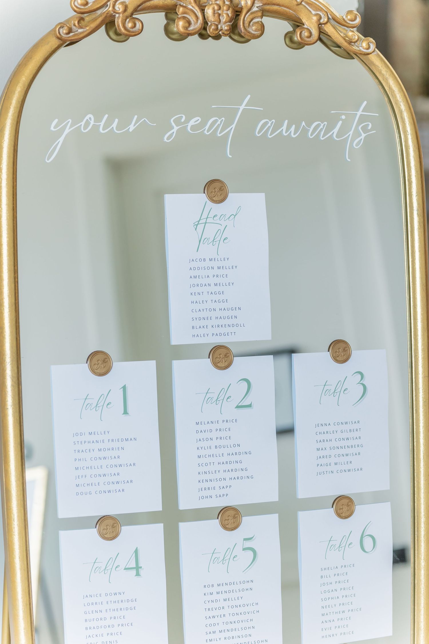 Elegant gold-framed mirror displays wedding seating chart cards with table numbers arranged in organized rows.