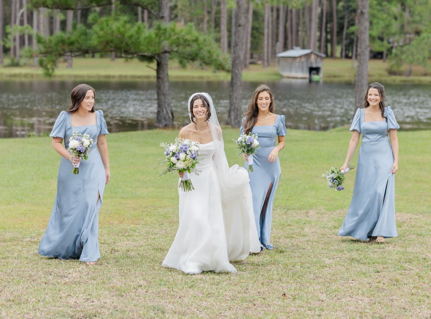 A group in light blue dresses and white gown gather playfully on a grassy lawn for outdoor wedding photos.