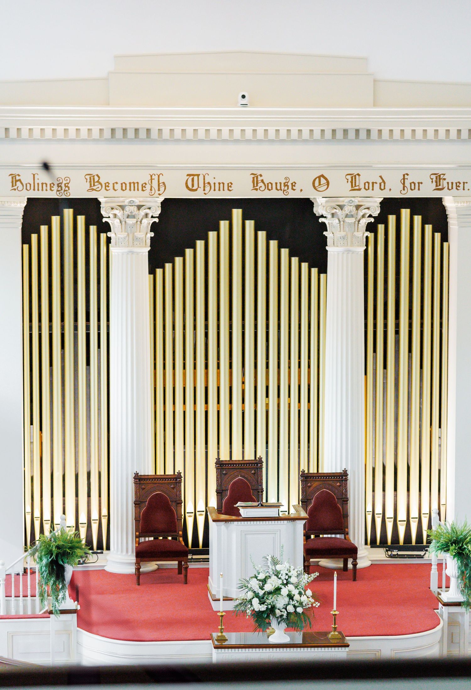 Grand church organ with gold pipes, white columns, red carpet altar, and wooden chairs in an elegant sanctuary.