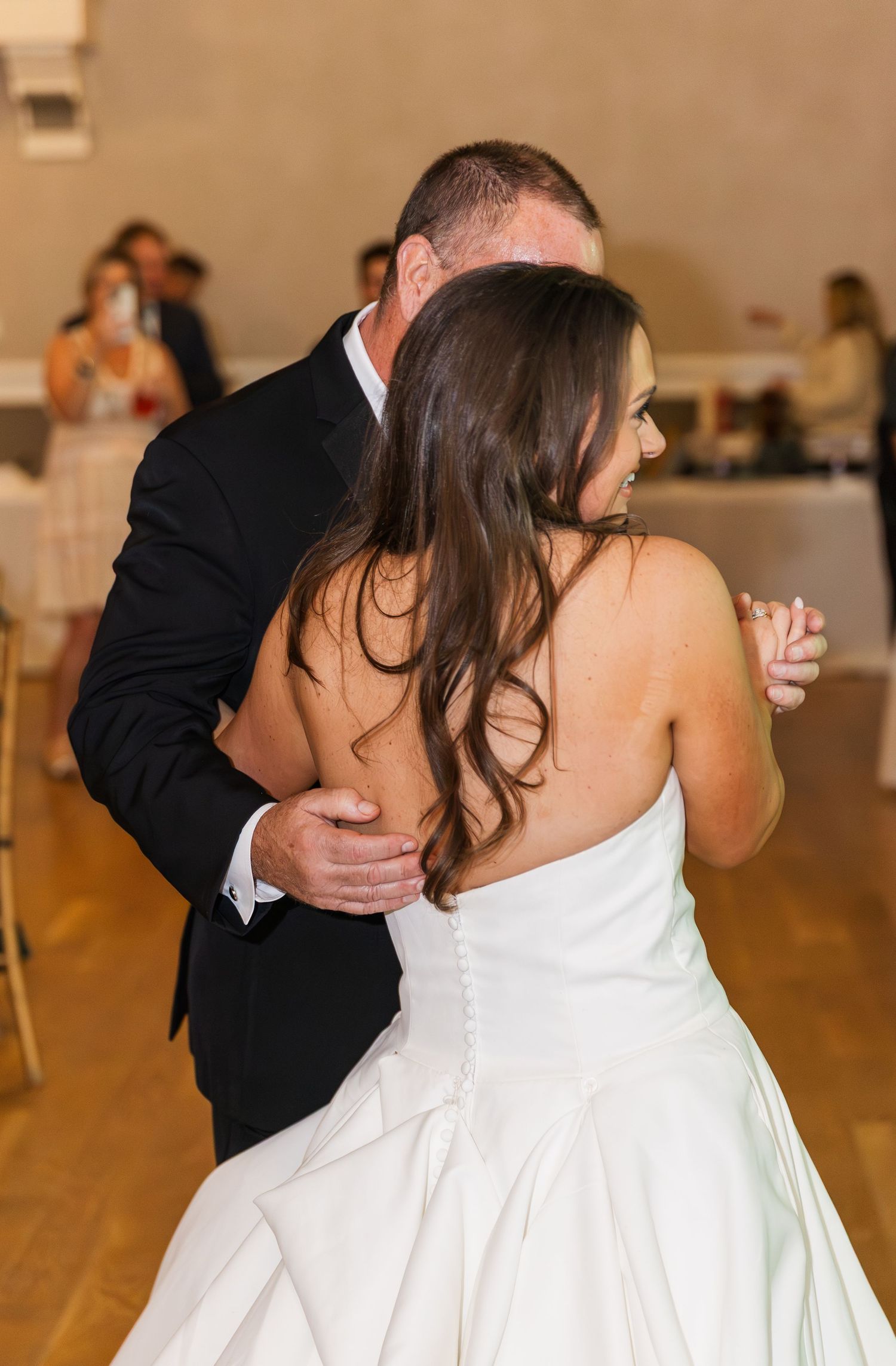 Bride in strapless white gown dances with man in black tuxedo at wedding reception.