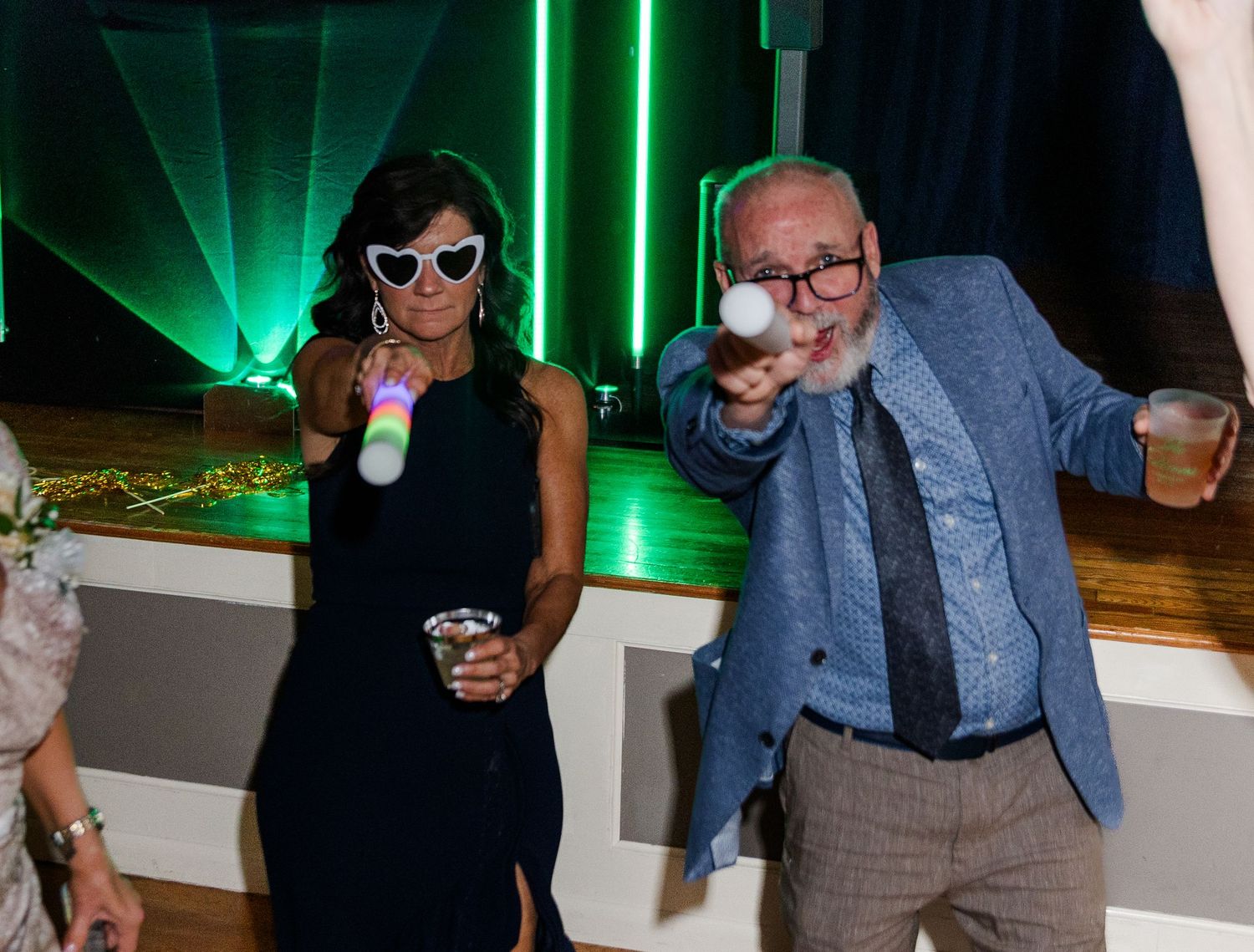 Two party guests wearing fun accessories dance at an evening event with green laser lights in the background.
