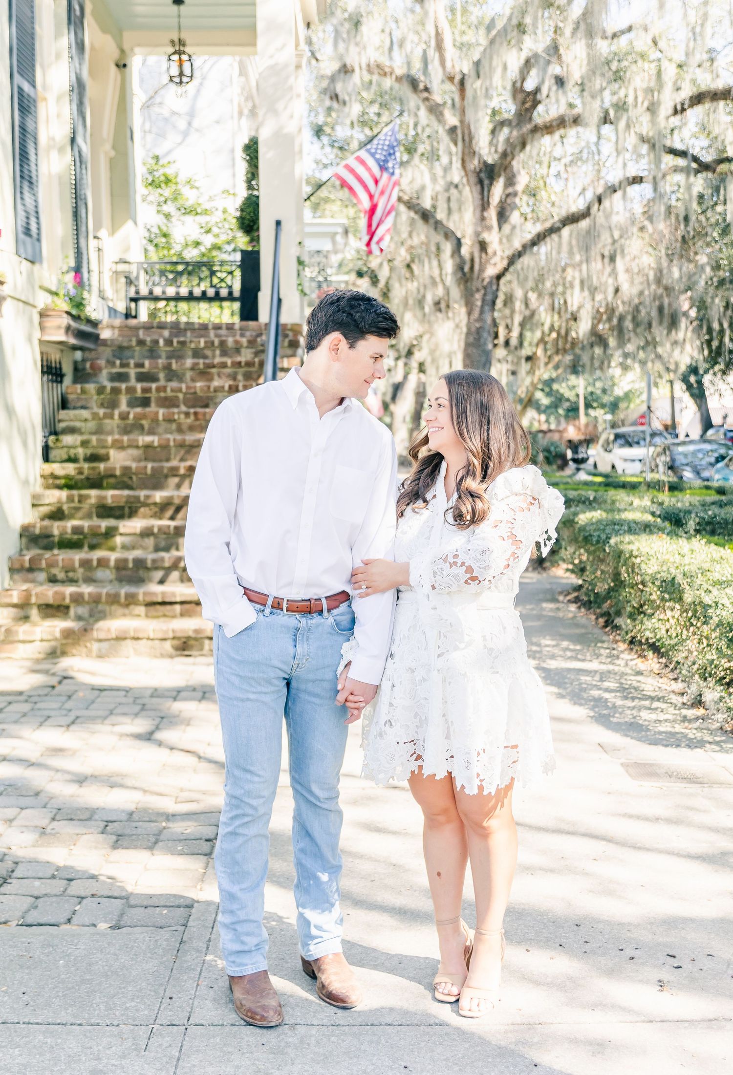 Romantic couple stands on cobblestone path under Spanish moss draped trees with American flag in background.