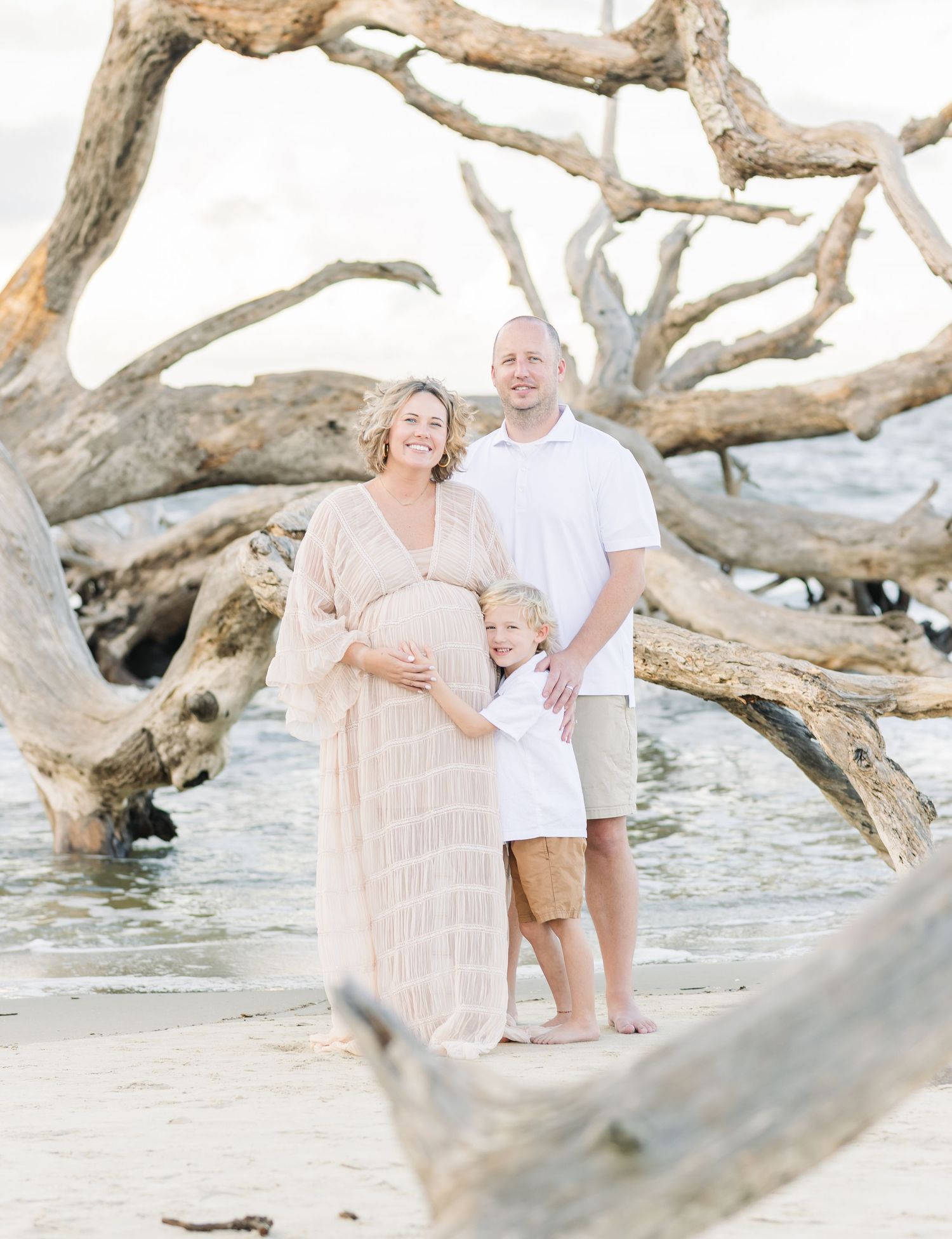 A family poses together on a beach with driftwood, wearing light colored clothing.