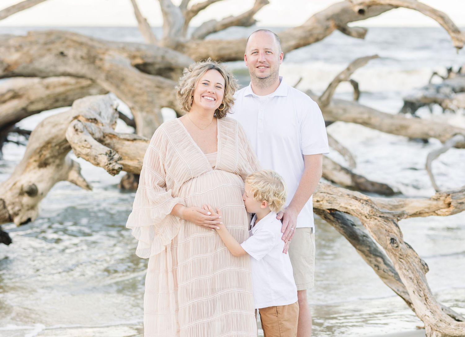 Family beach photoshoot among driftwood on sandy shore during sunset in neutral colors.